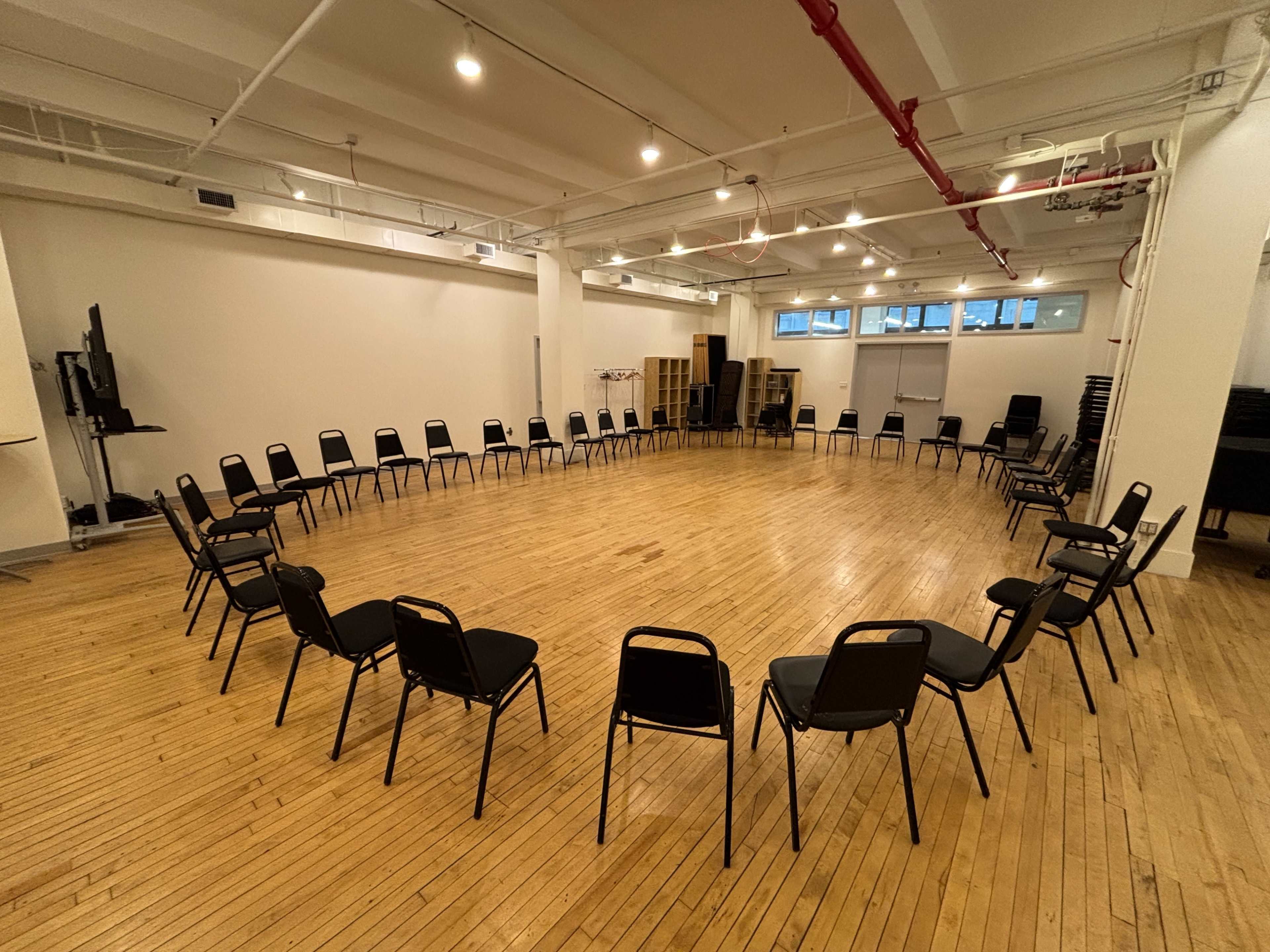The image shows a large, empty room with wooden floors arranged in a circle of black chairs.