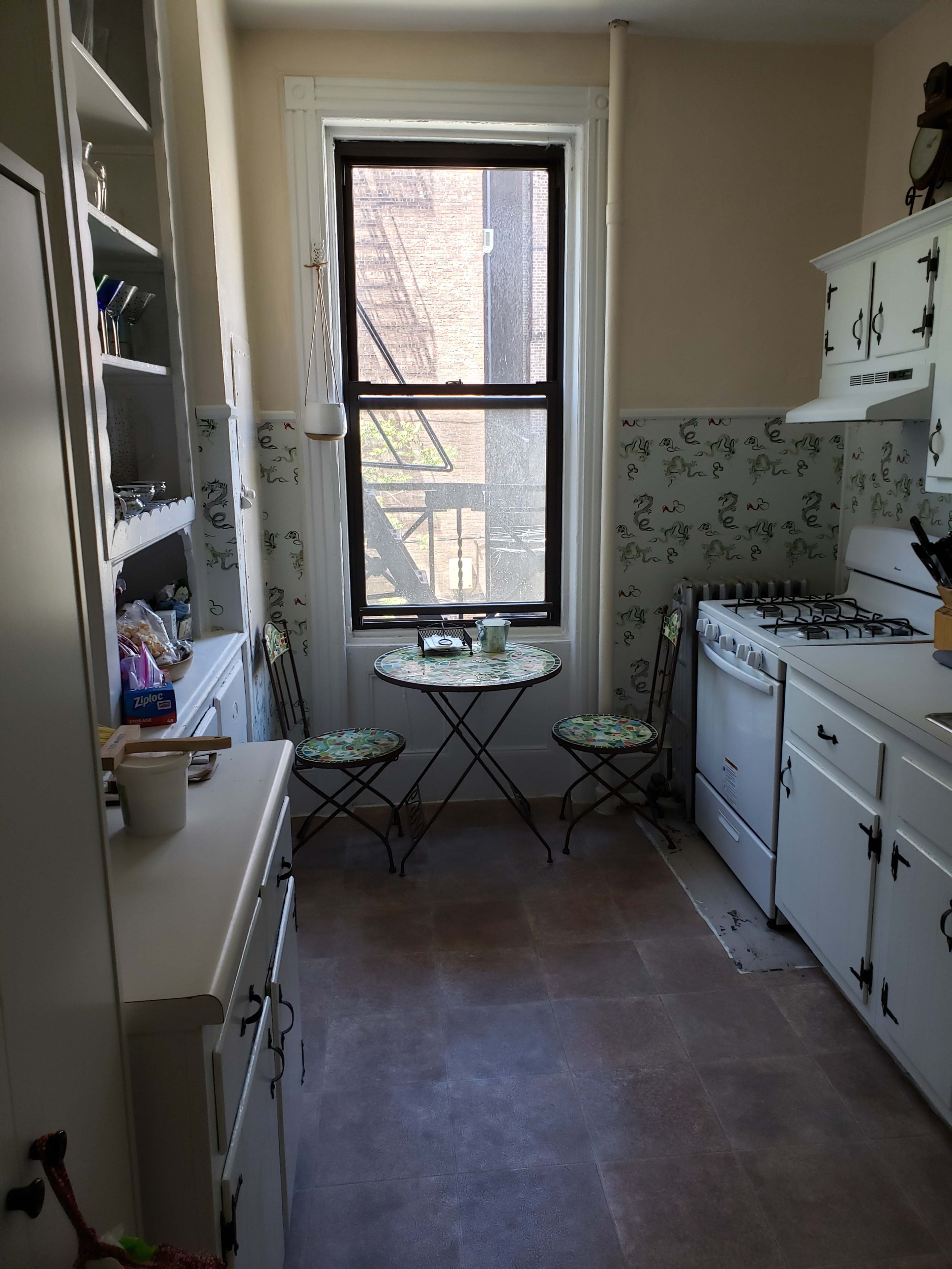 A small kitchen features a gas stove, white cabinets, and a compact dining table with two chairs near a window overlooking a fire escape.