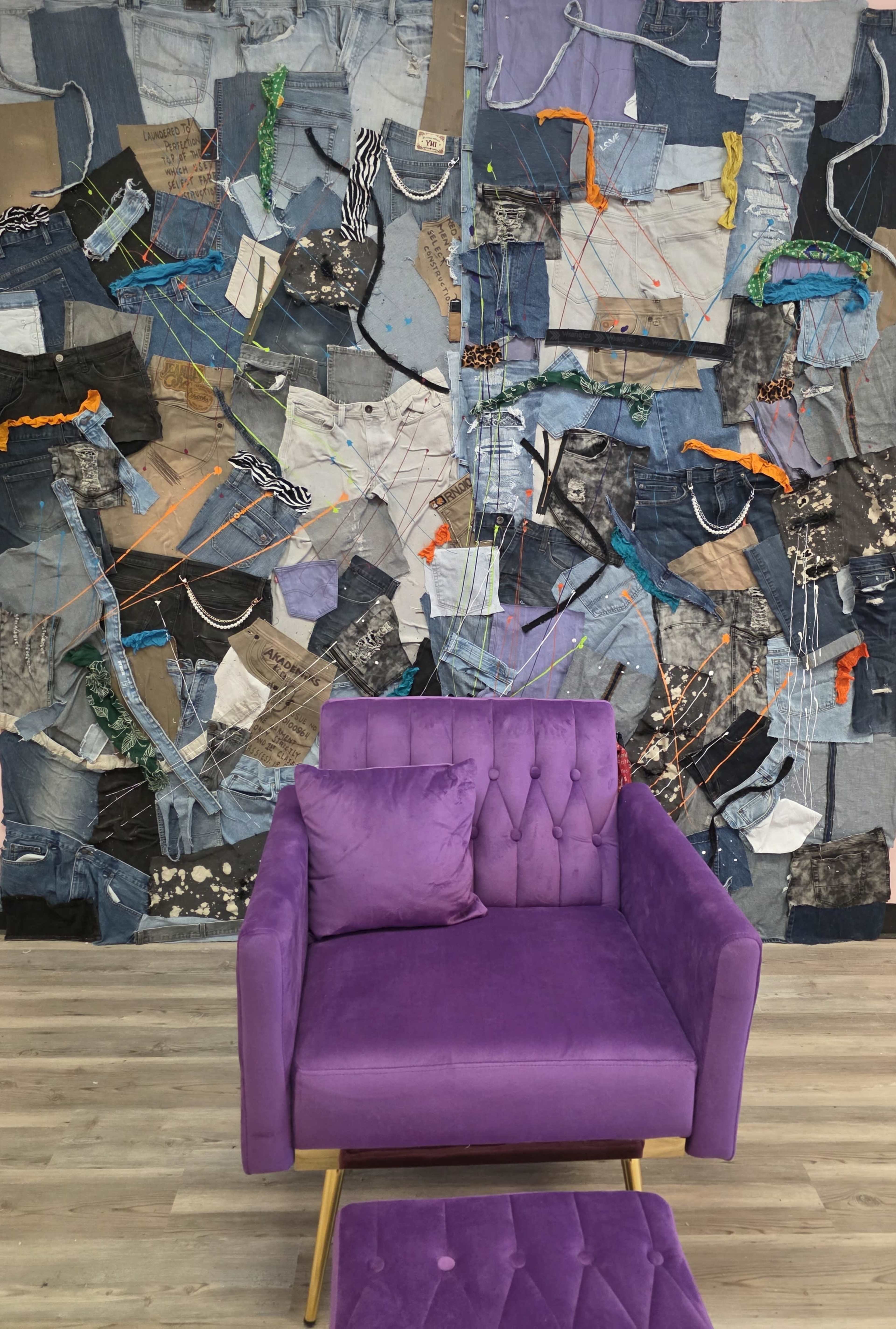 A purple velvet chair and ottoman are positioned in front of a large collage made of various pieces of denim and fabric, decorated with colorful threads.