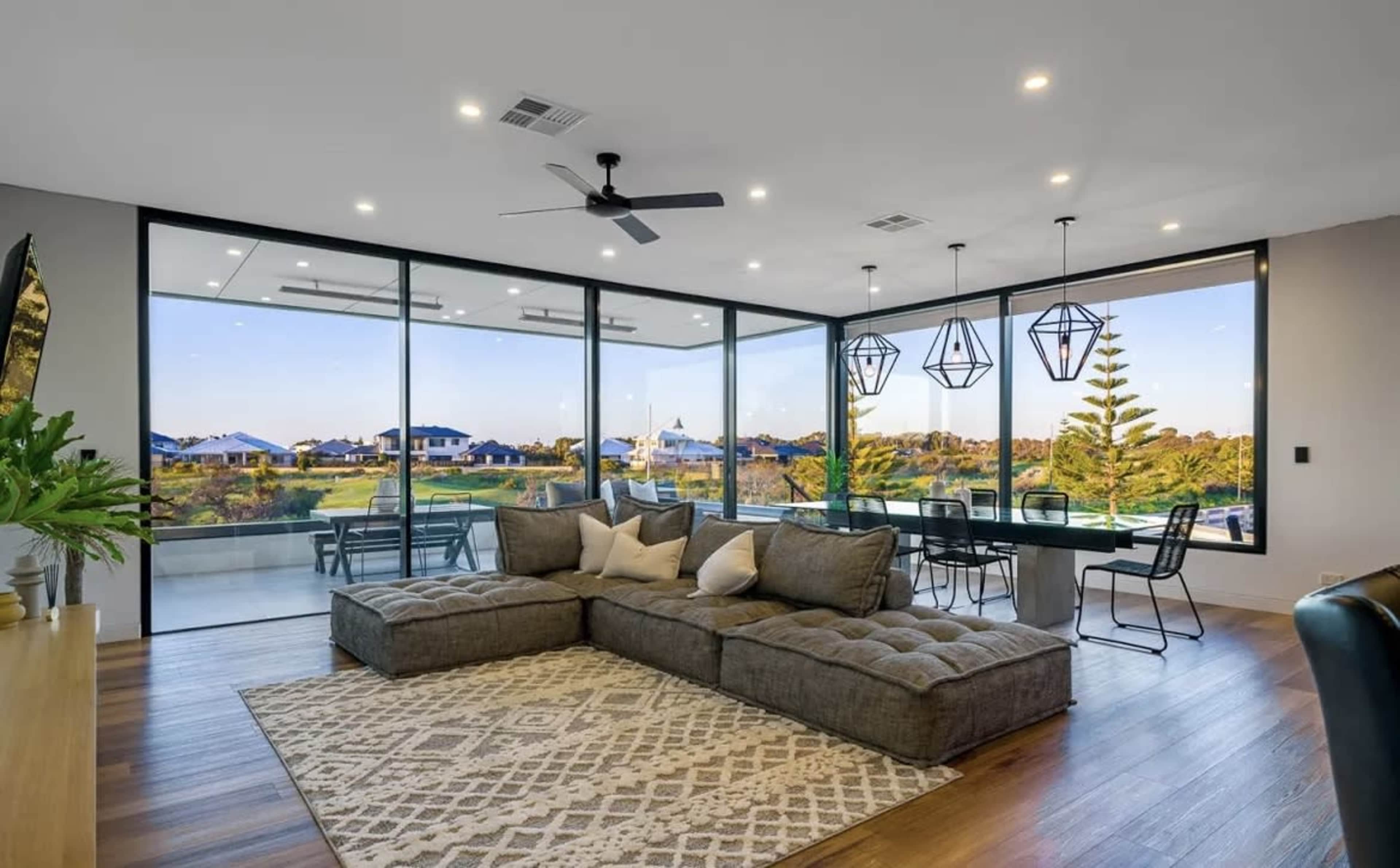 The image shows a modern living room with a large sectional sofa, a patterned rug, and glass walls that offer a view of a landscaped outdoor area.