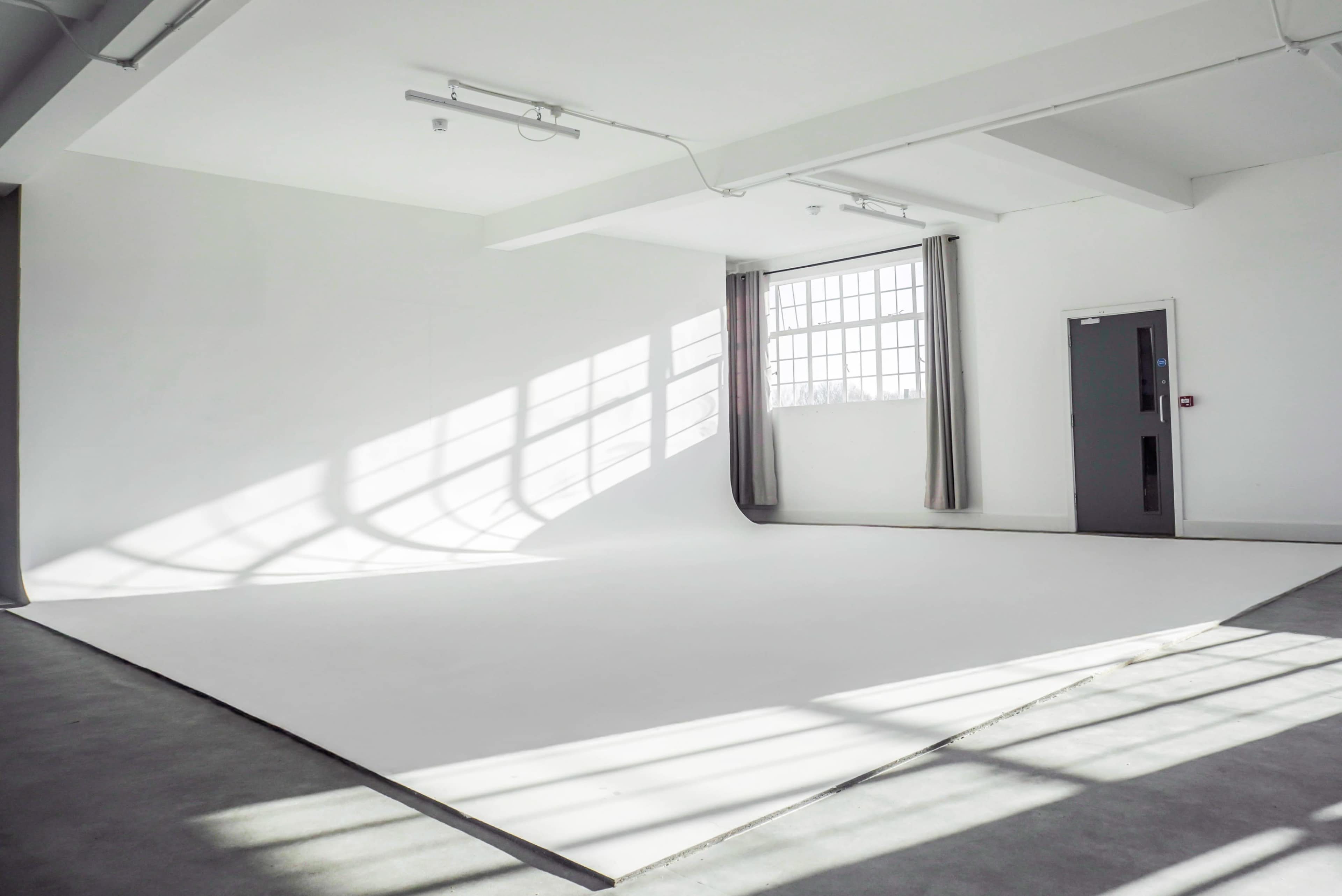 The image shows a bright, empty studio space with white walls, a large window, and soft shadows on the floor.