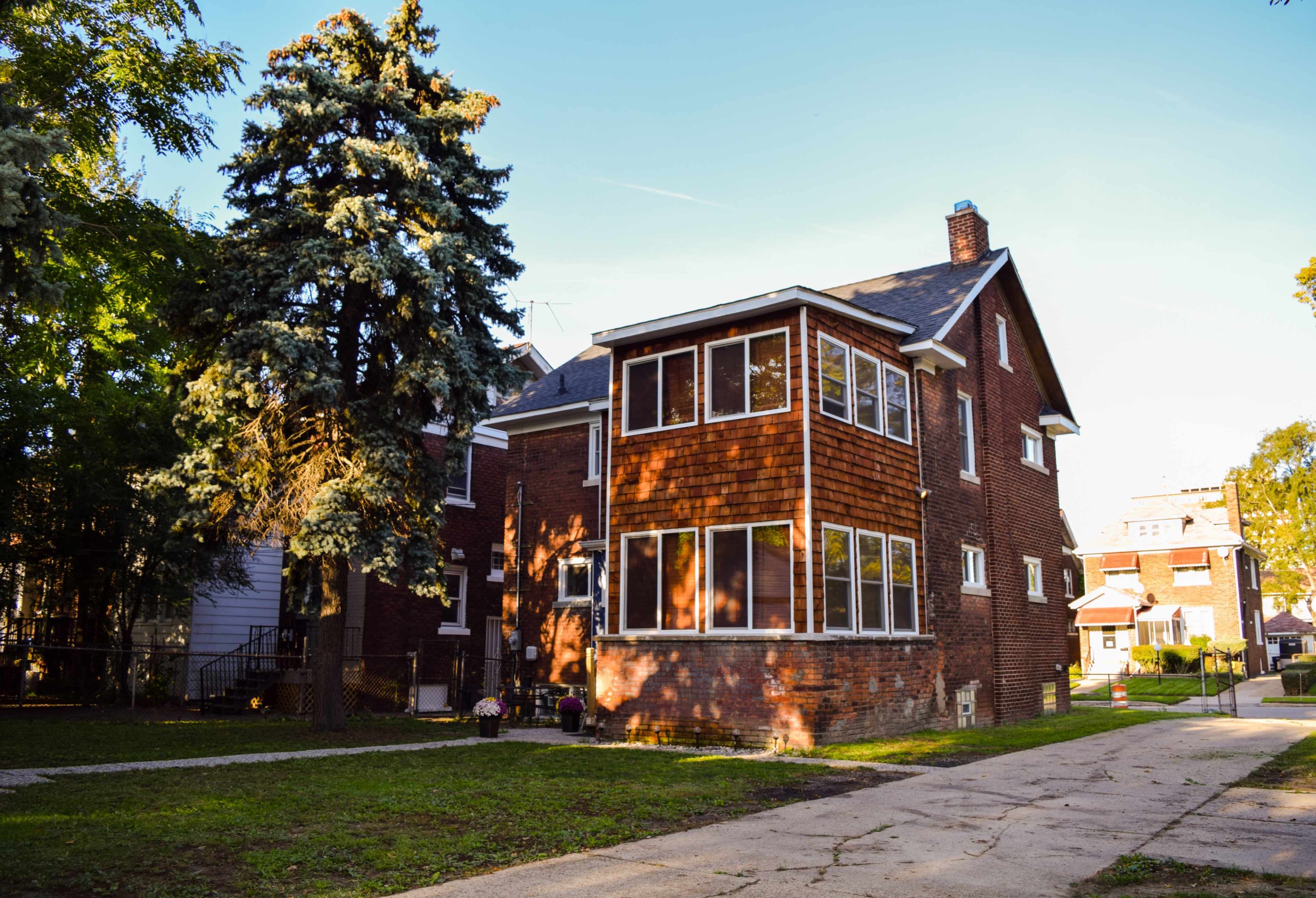 A two-story brick house features a large sunroom with numerous windows, surrounded by grassy areas and trees.