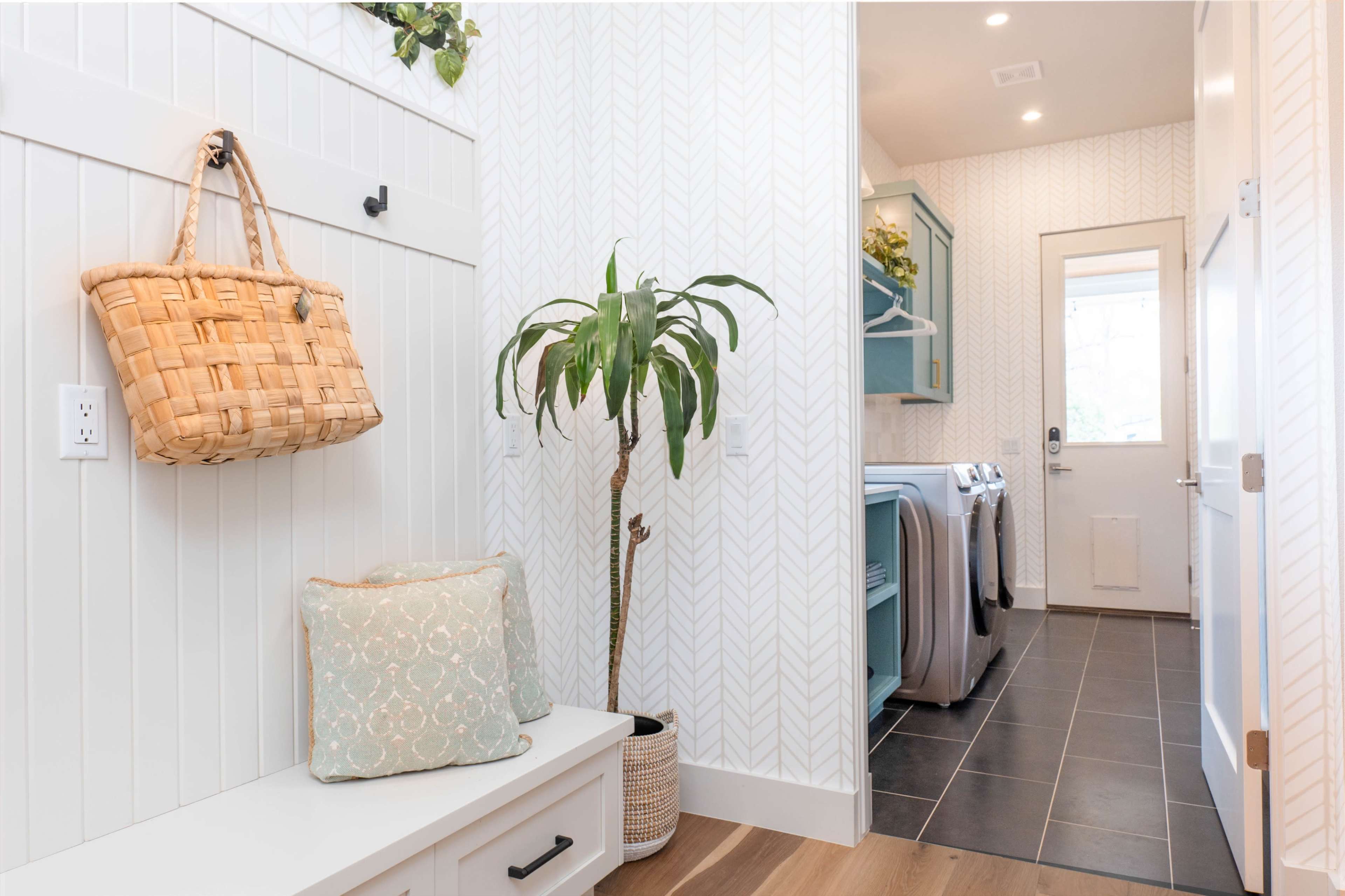 The image shows a hallway with a bench, a decorative basket hanging on the wall, a potted plant, and a doorway leading to a laundry area.