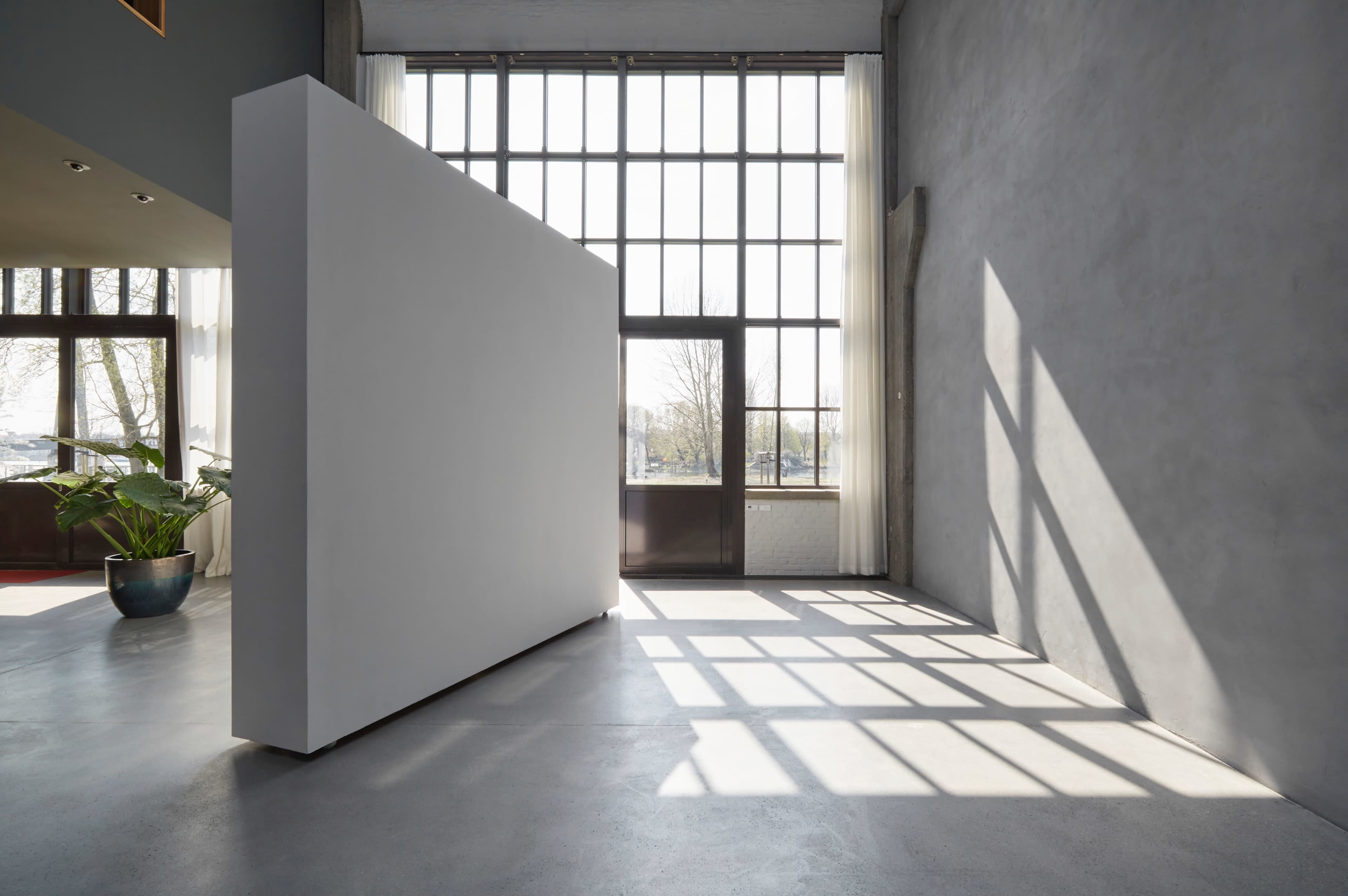 A large white partition stands in a well-lit room with tall windows, casting shadows on the concrete floor.