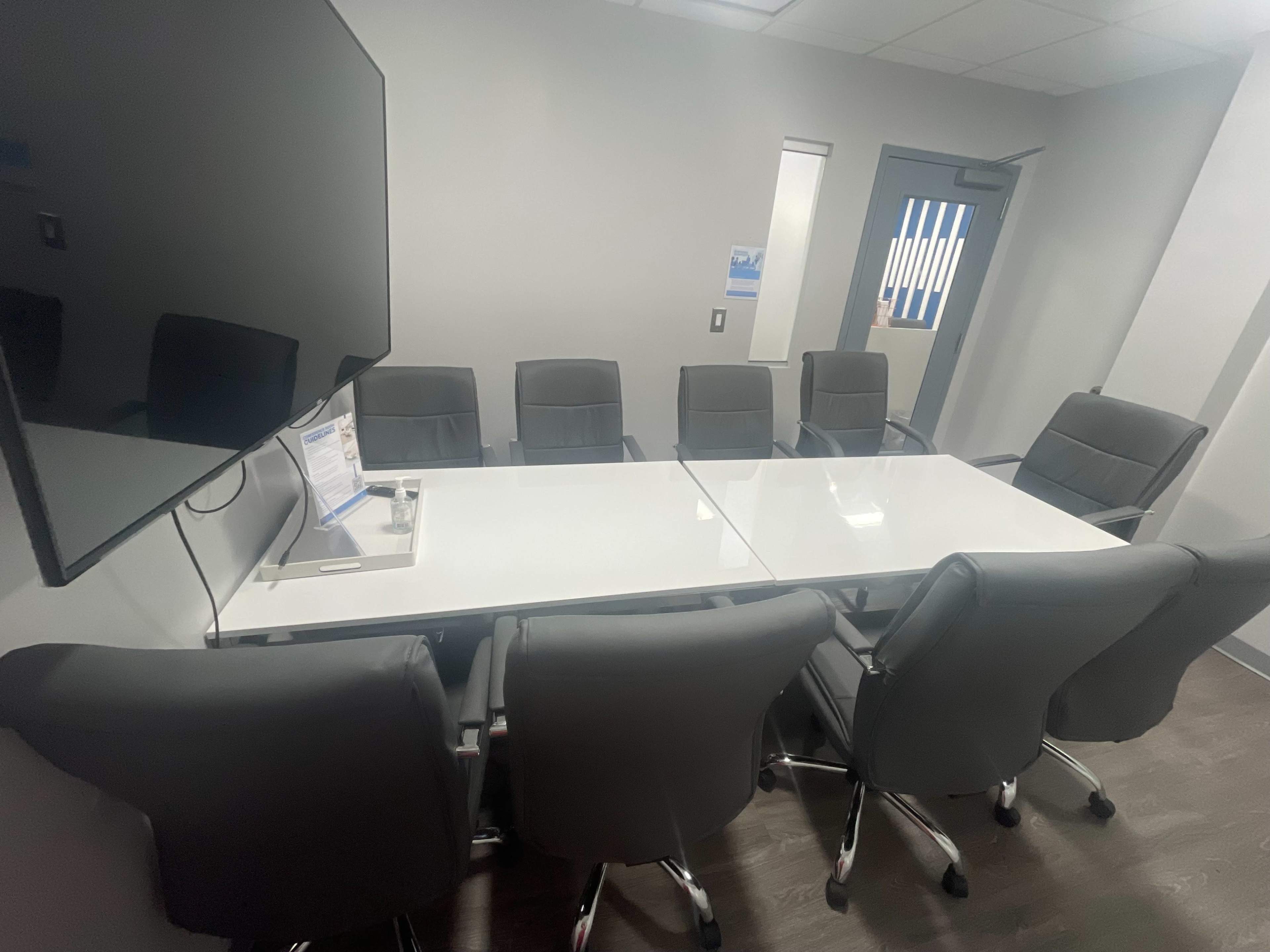 The image shows a conference room with a glass table surrounded by eight black leather chairs, and a television mounted on the wall.