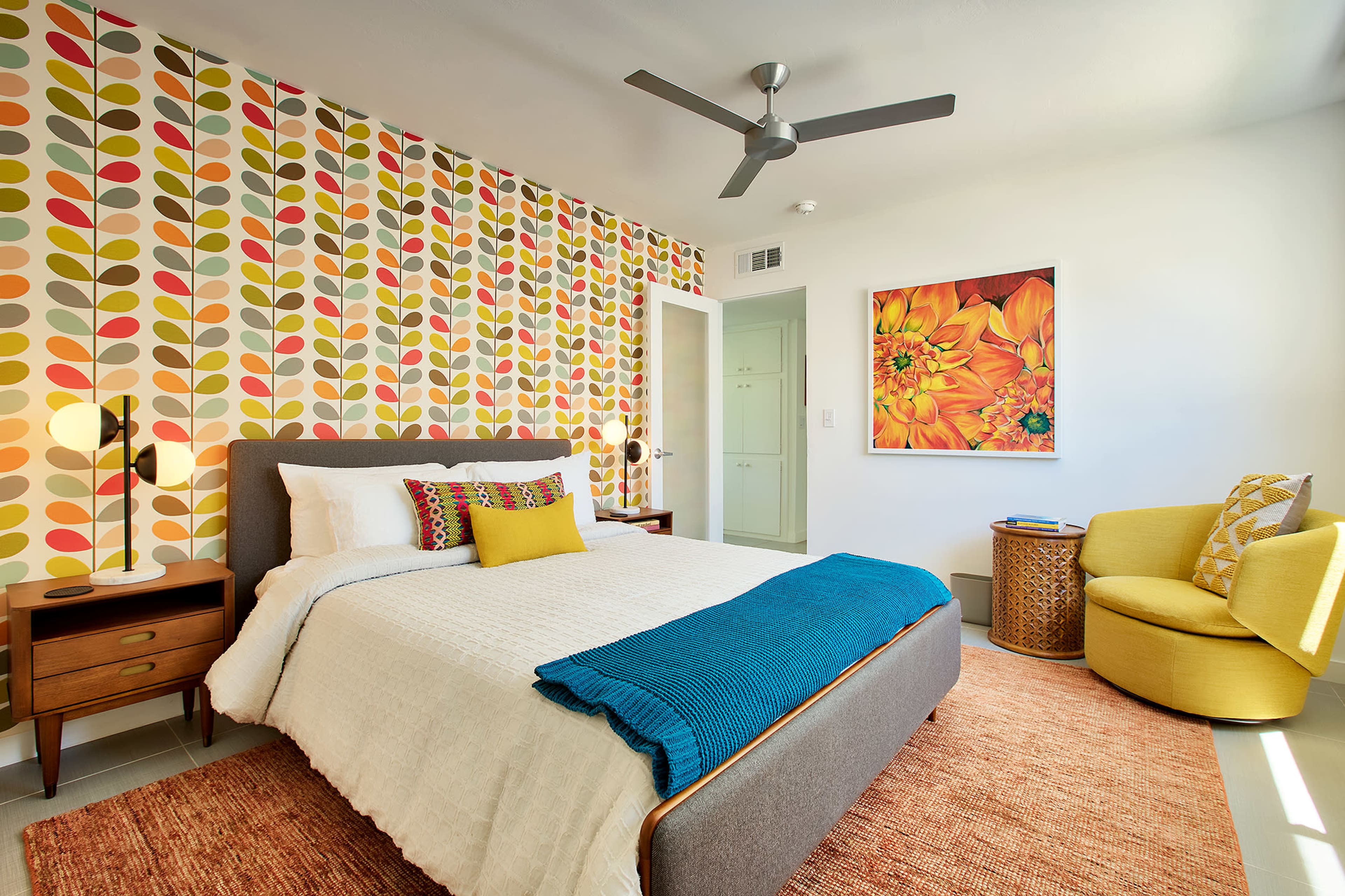A modern bedroom featuring a bed with a colorful accent wall adorned with leaf patterns, a yellow accent chair, and a vibrant floral artwork.
