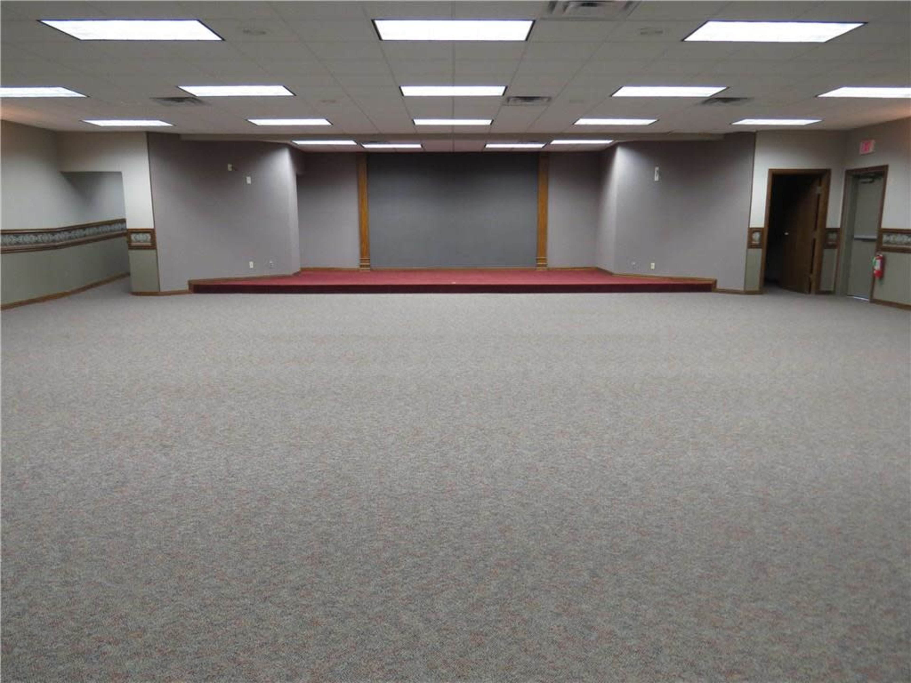 A vacant room features a raised stage at one end and is carpeted throughout.