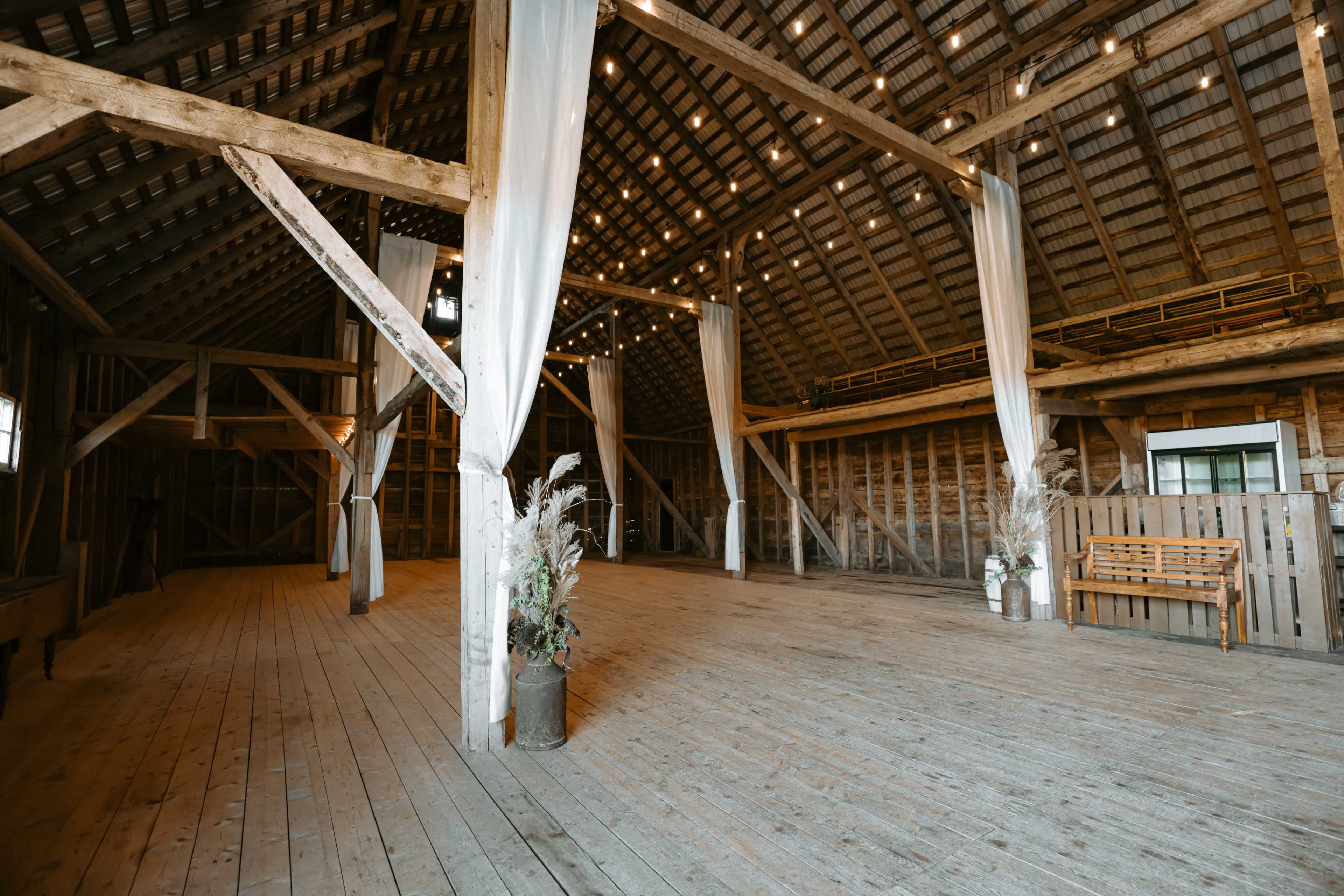 The image shows the interior of a spacious, rustic barn with wooden beams, a wooden floor, and draped fabric accents.