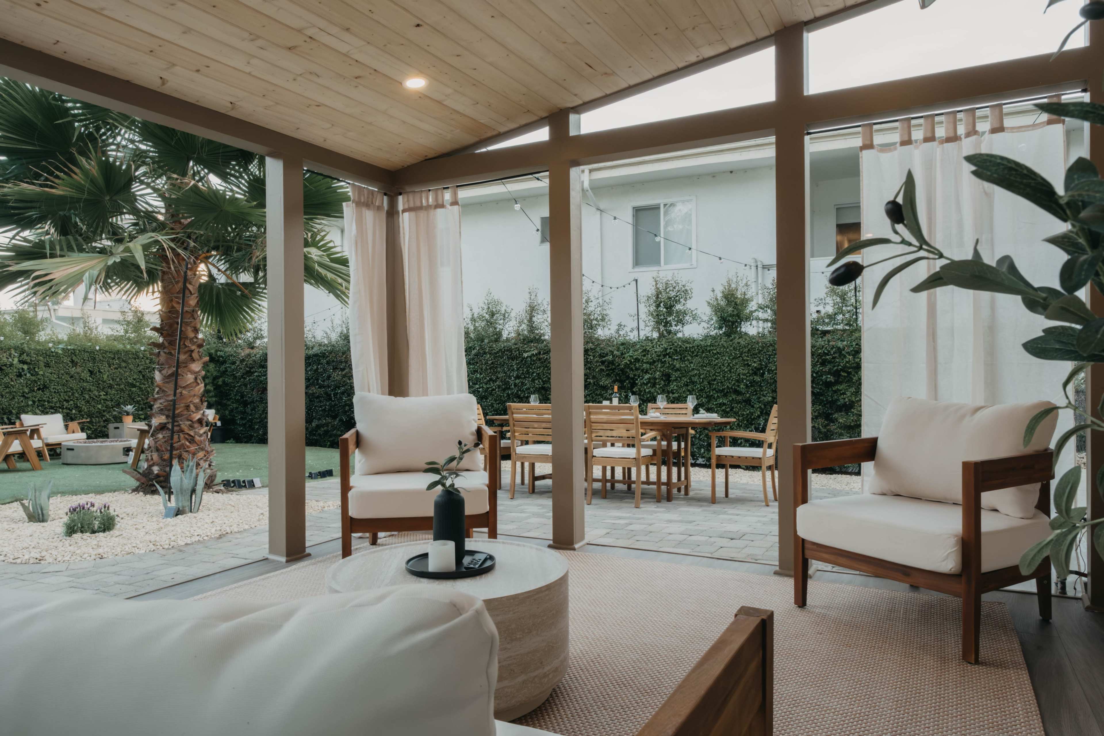 A covered patio area features two white armchairs and a round coffee table, overlooking a backyard with a dining set and palm tree.