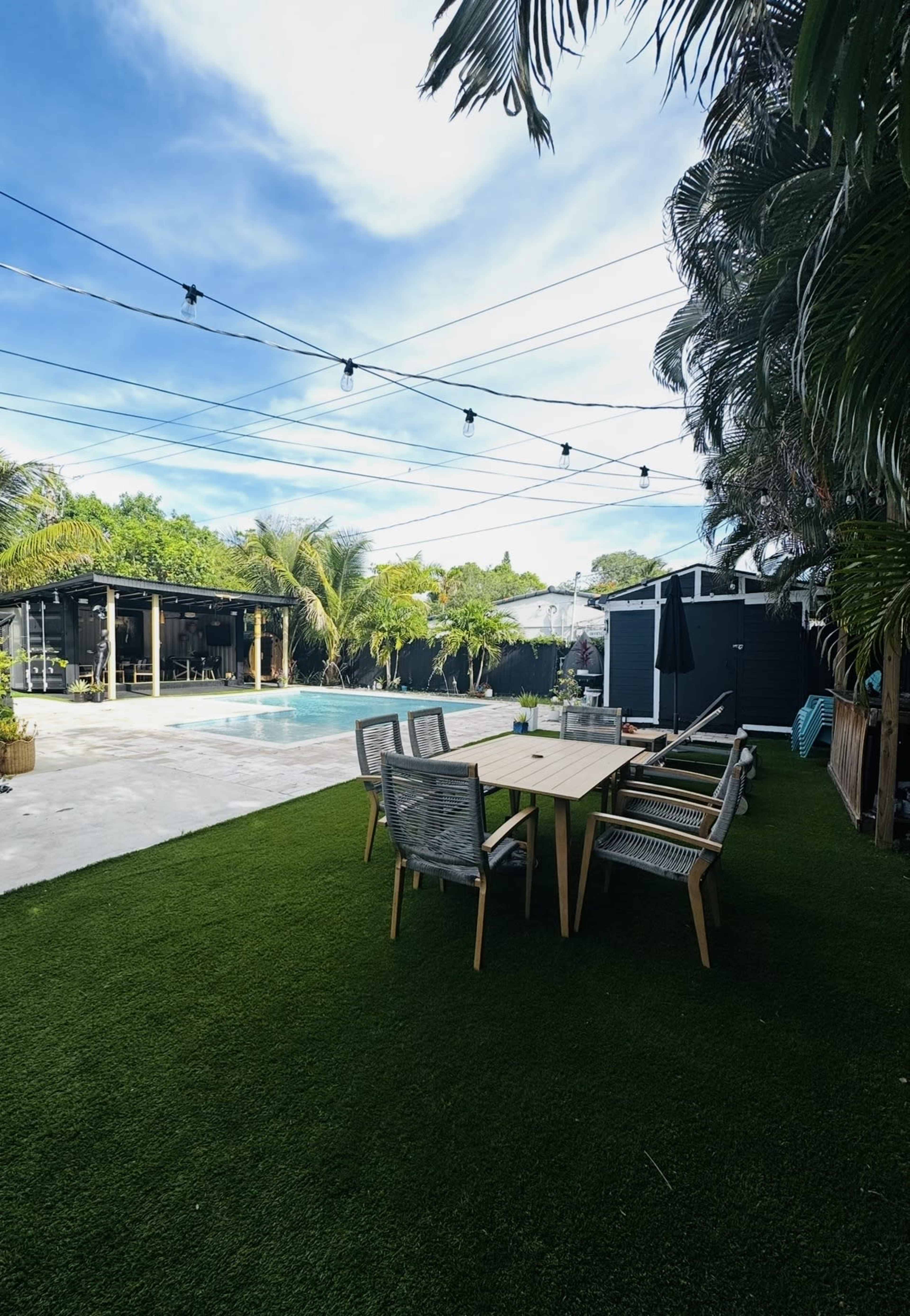 The image shows a backyard with a swimming pool, a dining table with chairs, and lush greenery surrounding the area under a clear blue sky.