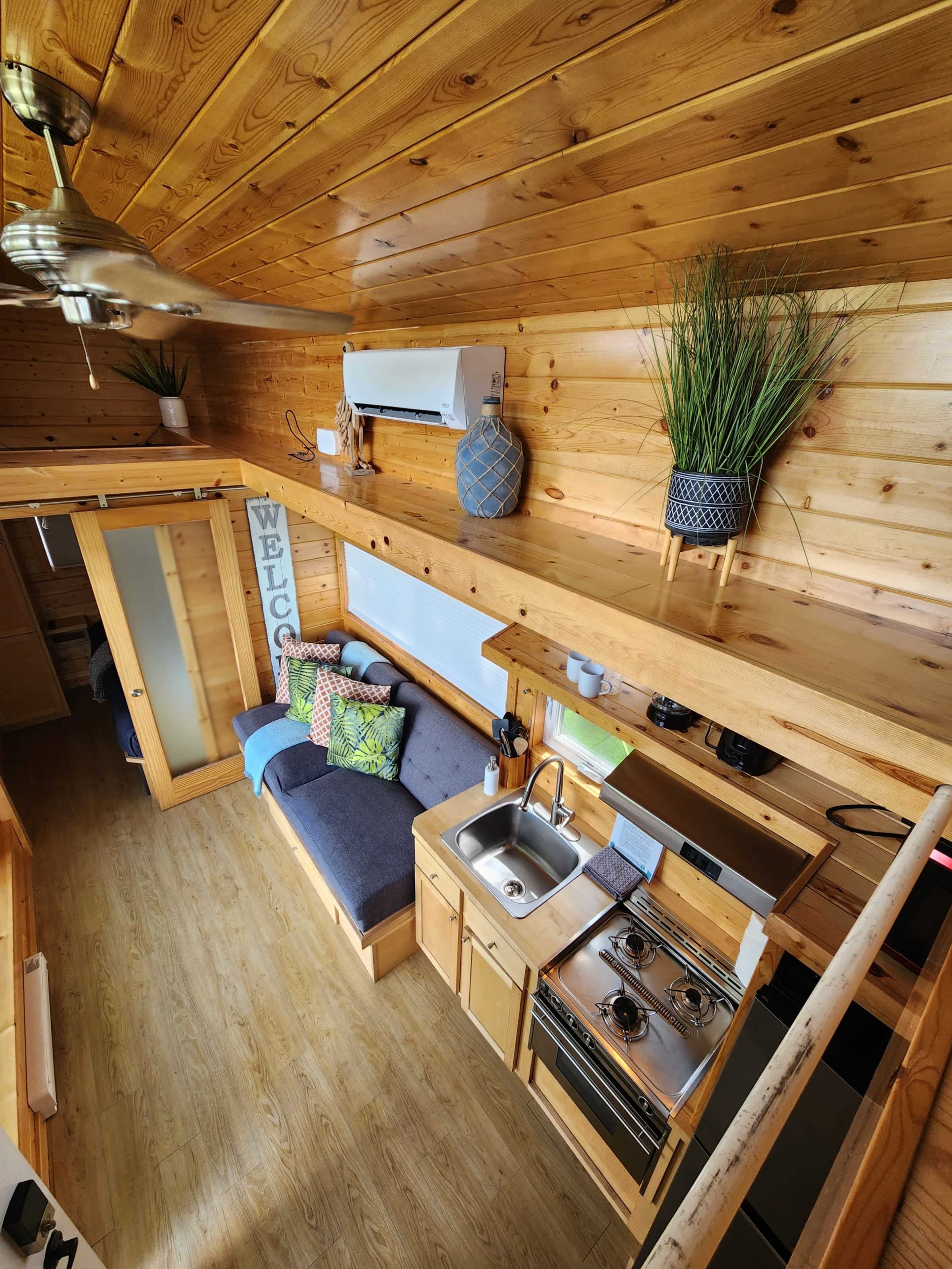 A compact wooden interior of a tiny home featuring a kitchenette, seating area, and decorative plants on shelves.