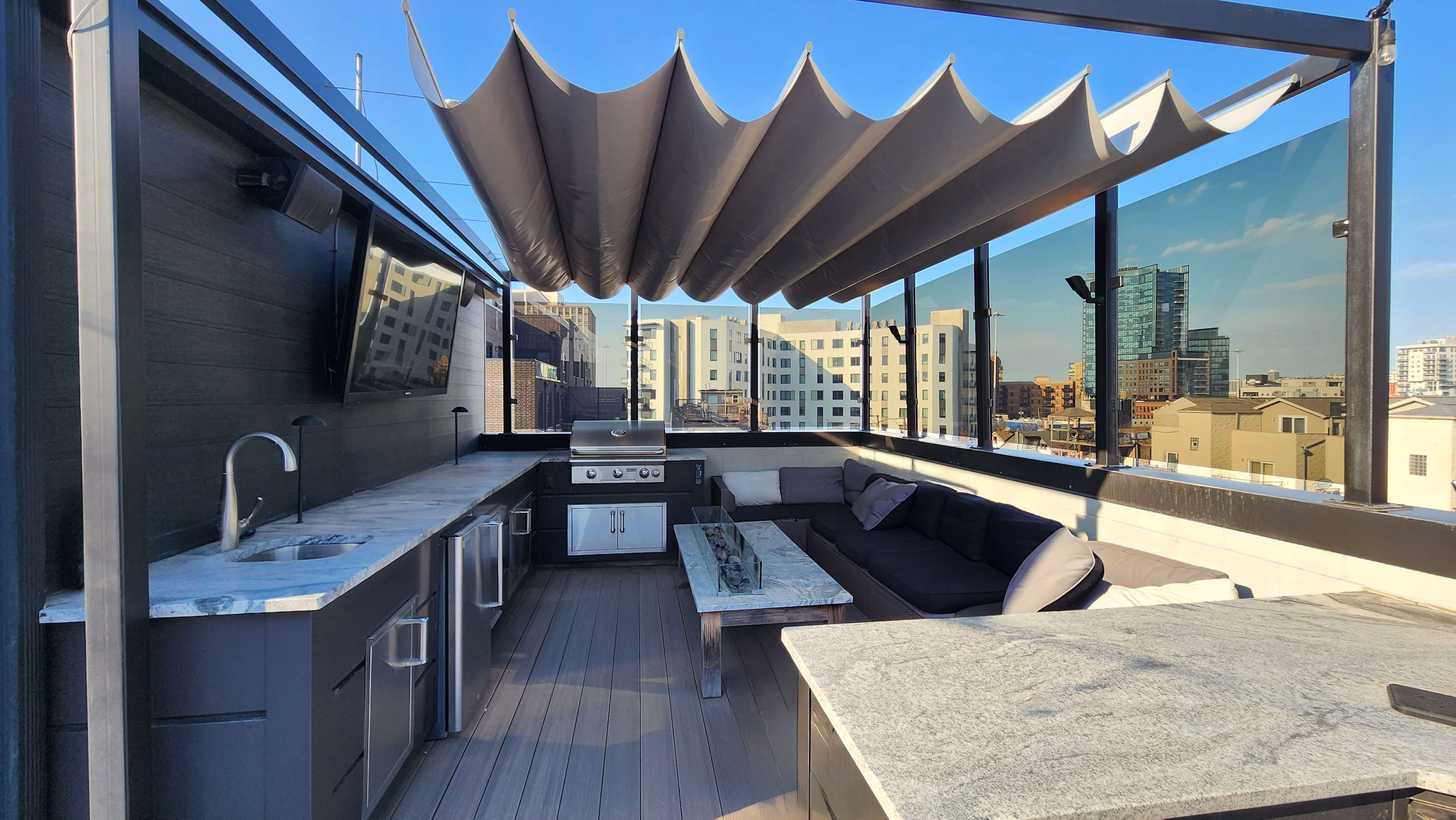 The image shows a rooftop terrace equipped with an outdoor kitchen, seating area, and a shaded canopy.