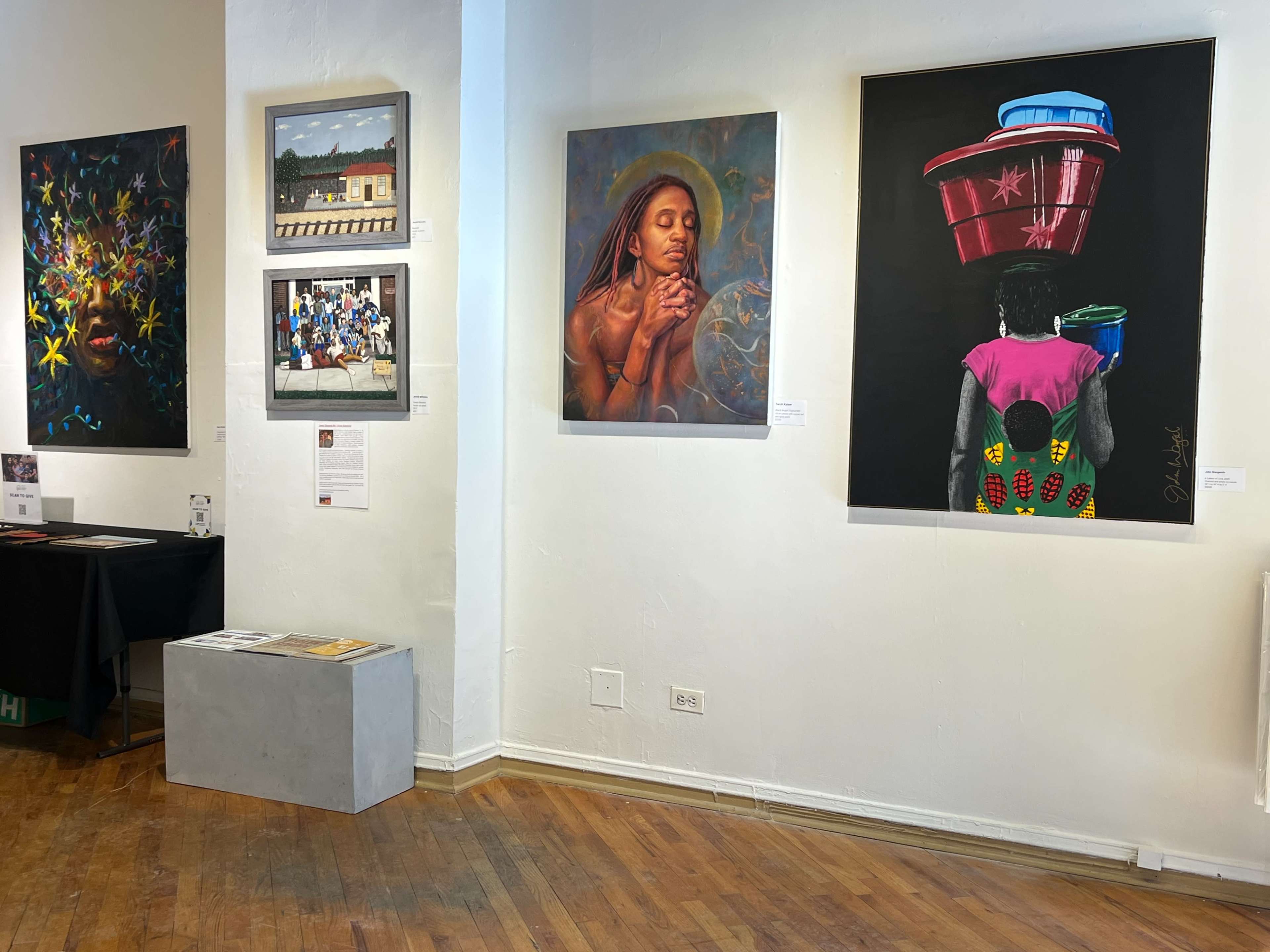 The image shows an exhibition space with various artworks displayed on the walls, including paintings and photographs, alongside a gray bench on the wooden floor.