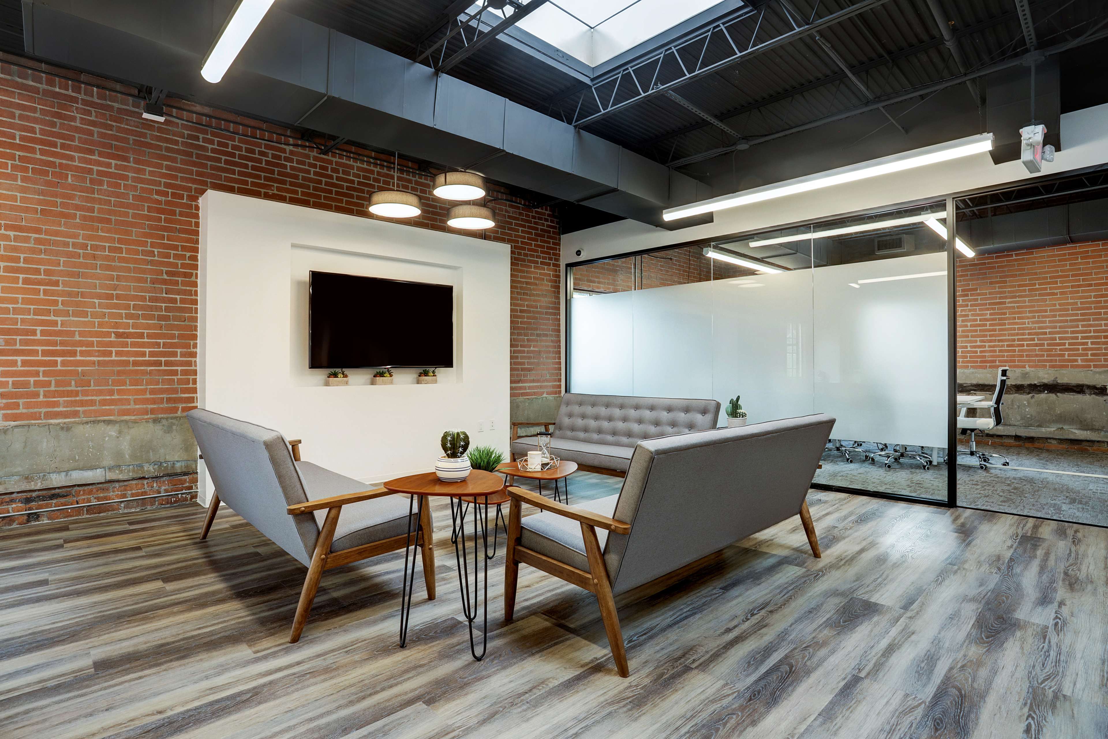 A modern office lounge area with gray sofas, a wooden coffee table, and a glass-walled meeting room in the background.
