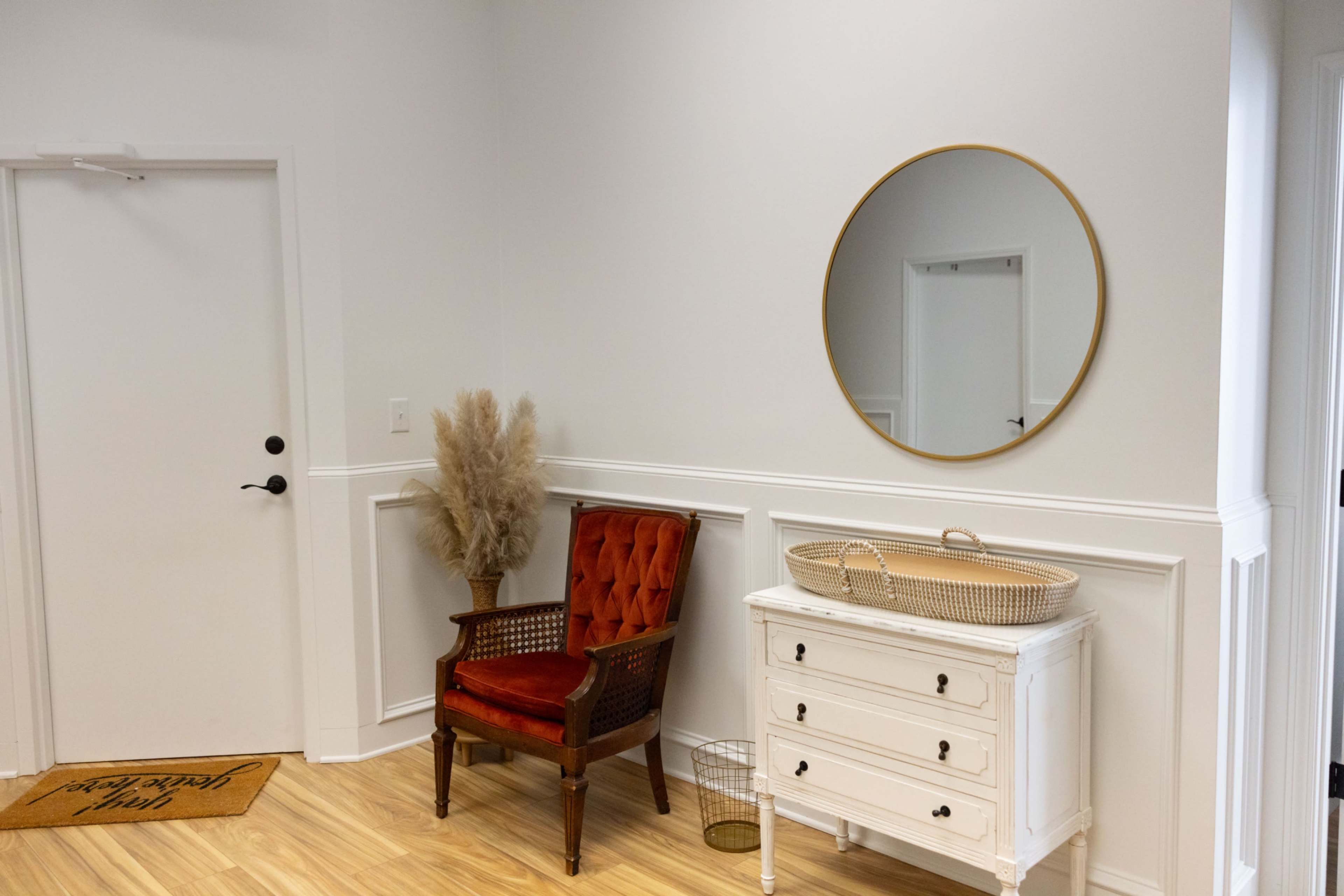 A red upholstered chair sits beside a white dresser and a round mirror on a light-colored wall.