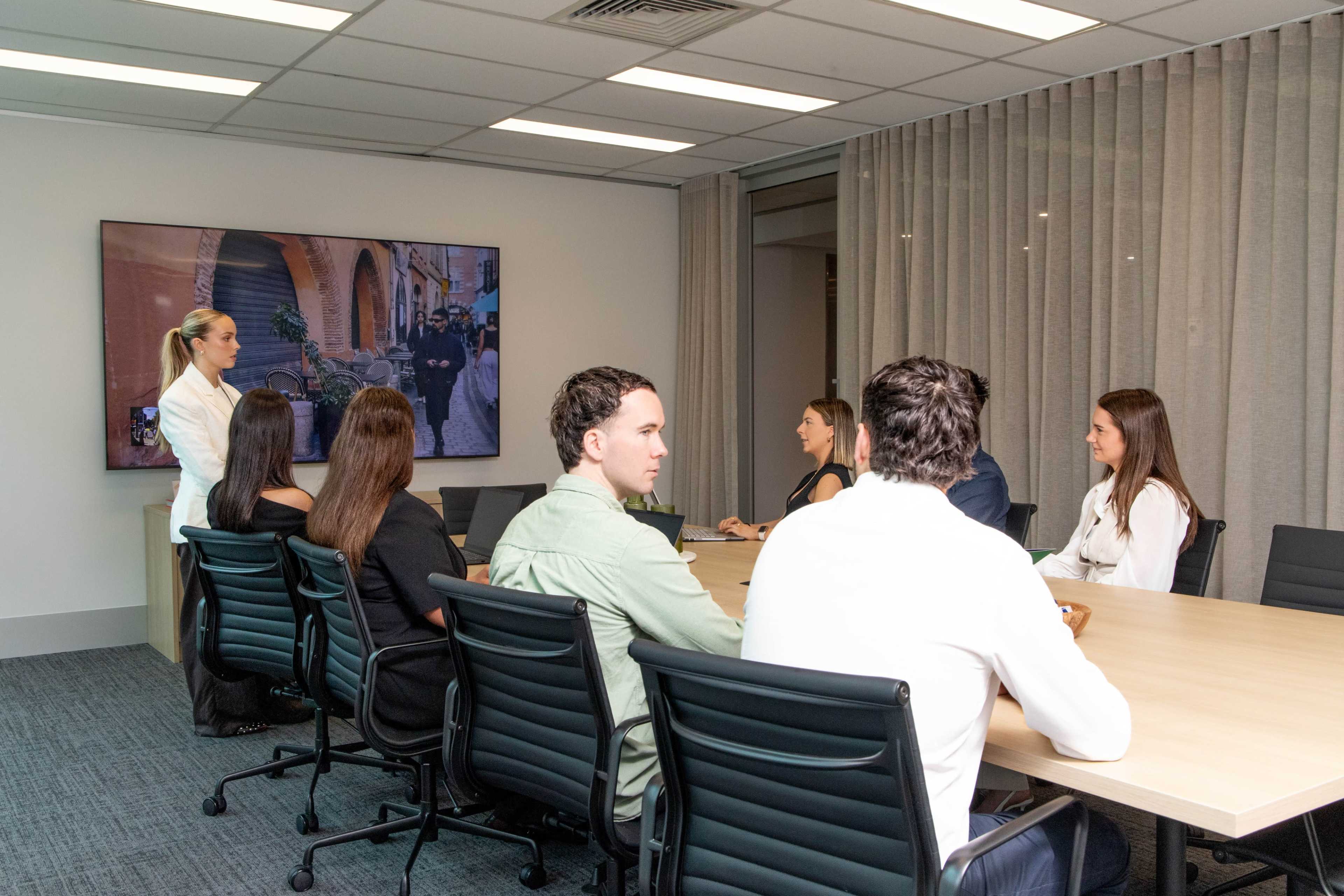 A group of six individuals is seated around a long conference table, while a woman stands in front of a screen displaying an image of a street scene.