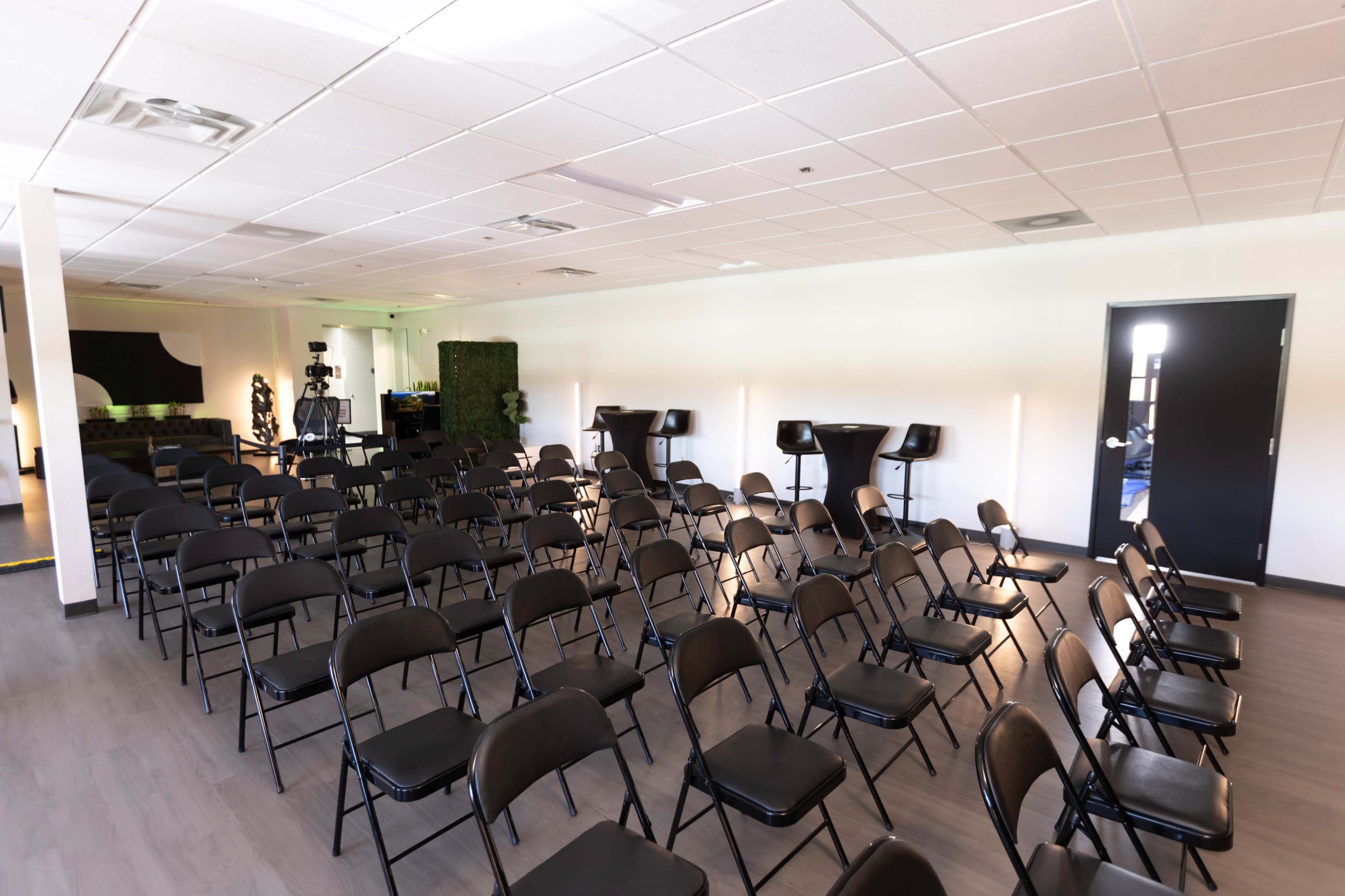 A neatly arranged room with rows of black folding chairs facing a presentation area and a few high-top tables along the sides.