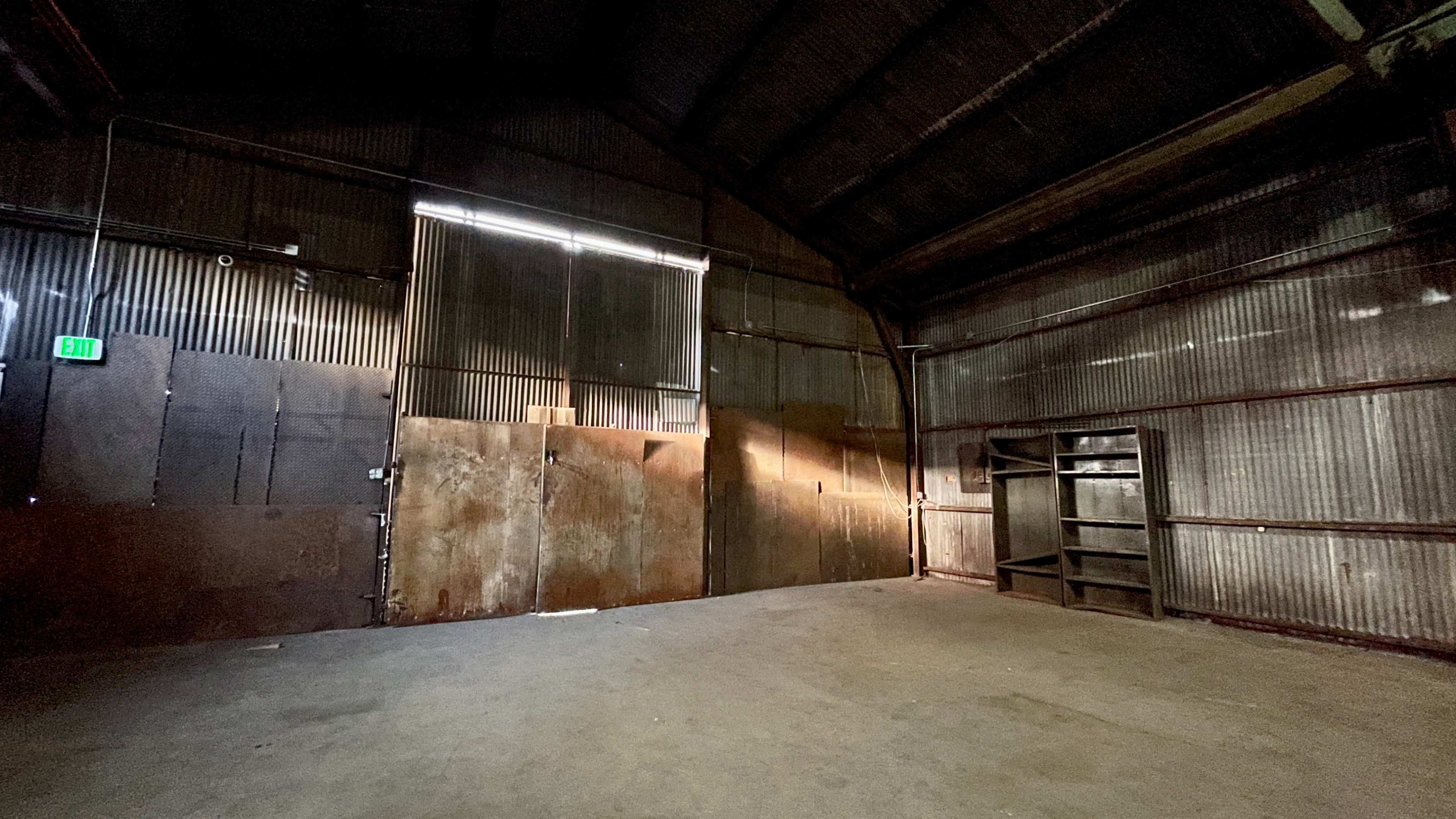 The interior of a large, empty warehouse with metal walls, sparse lighting, and a wooden shelving unit in one corner.