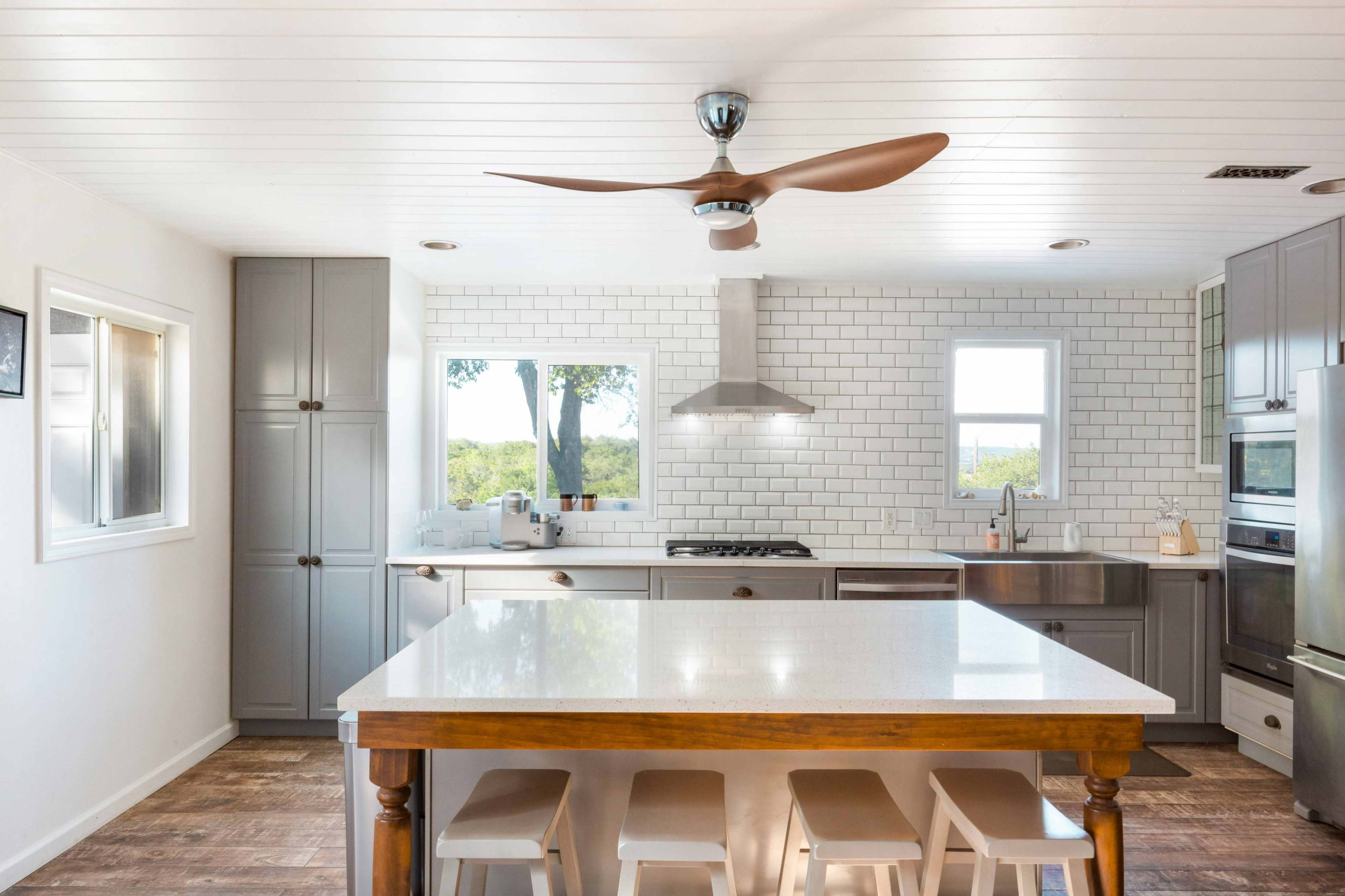 A modern kitchen features gray cabinetry, a large island with white countertops, and a stainless steel stove beneath a ceiling fan.
