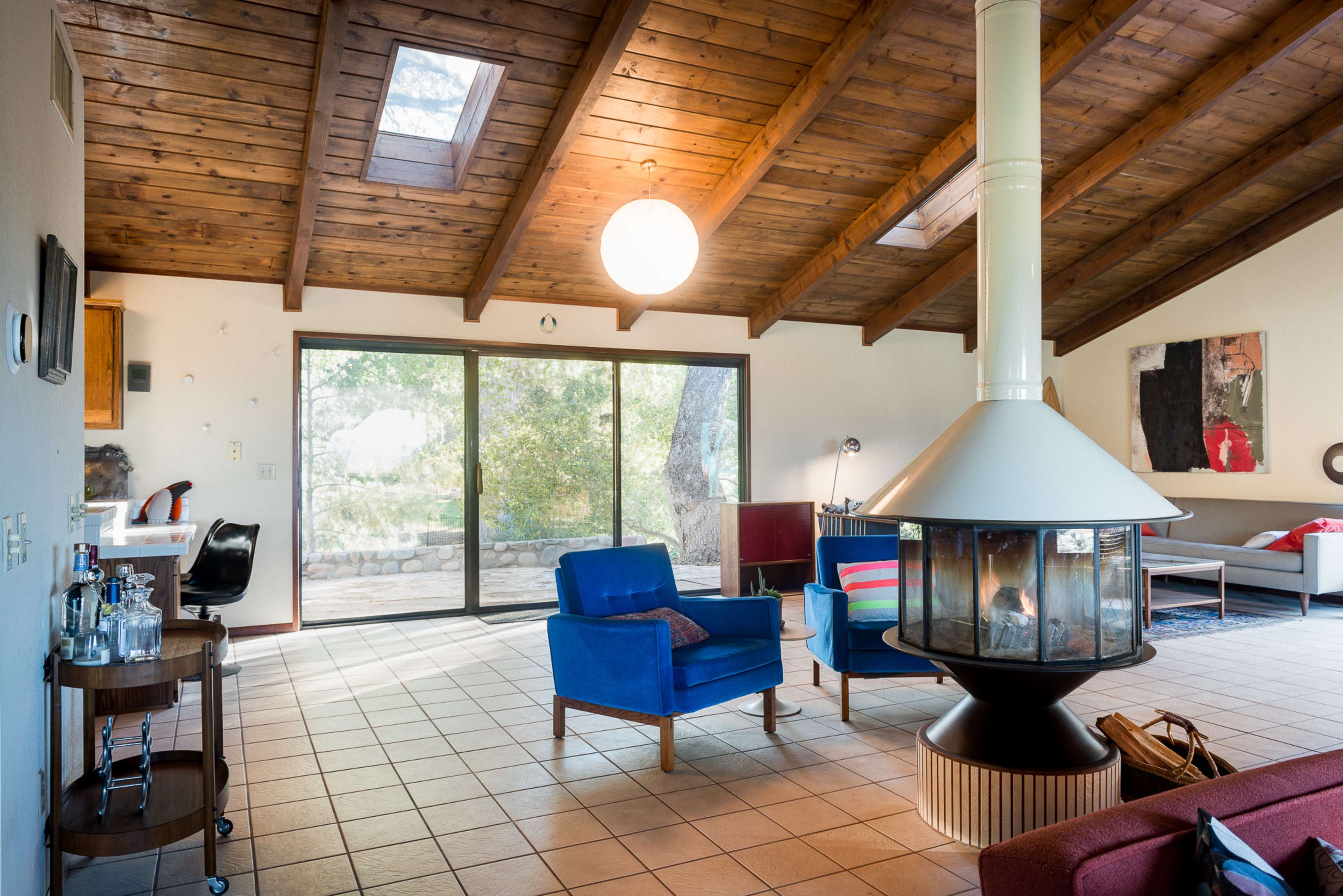 The image shows a spacious living room with a high wooden ceiling, large glass doors opening to an outdoor view, and a central round fireplace.