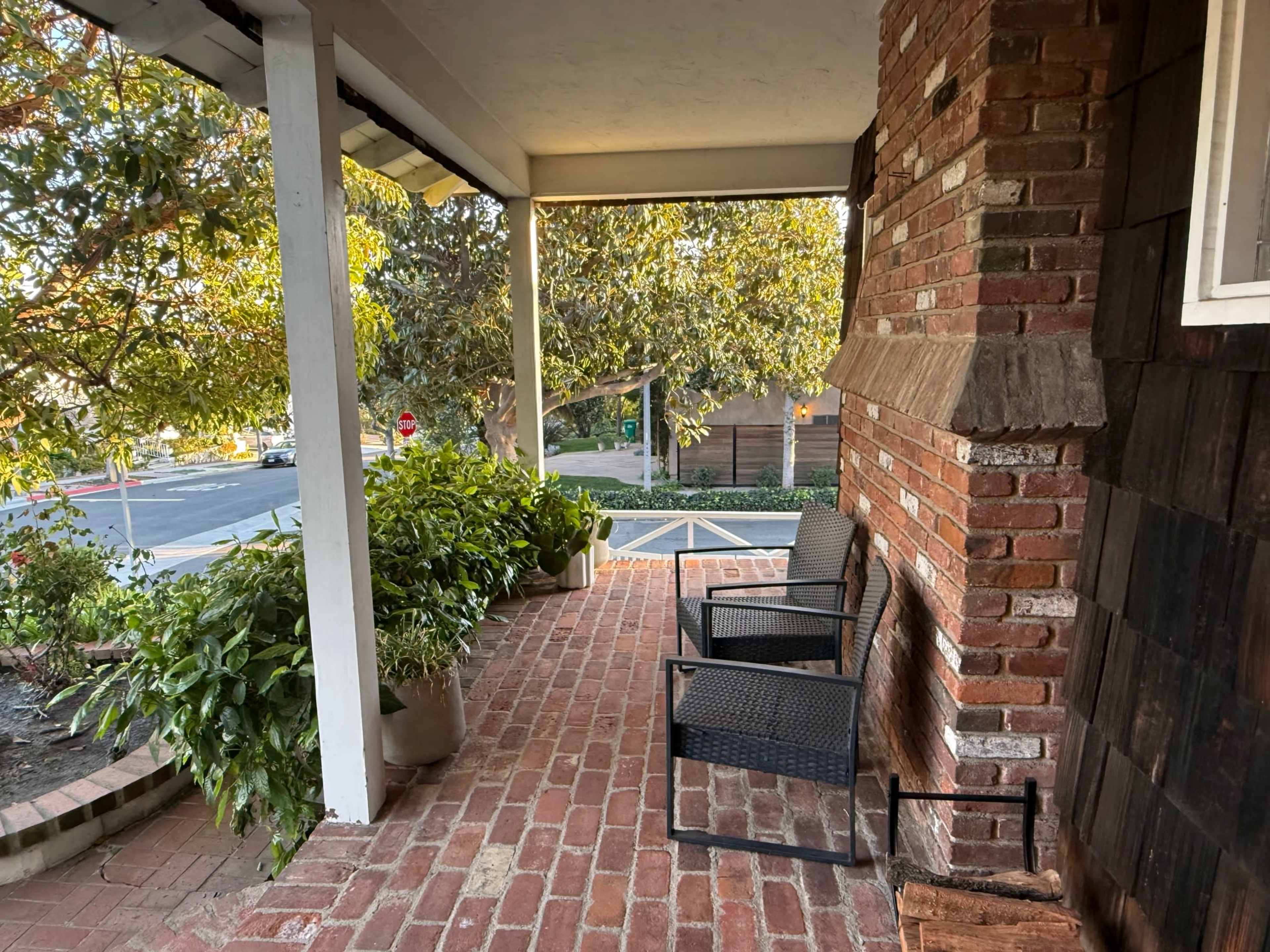 The image shows a porch with two black chairs positioned on a brick floor, surrounded by green plants, and a view of a street in the background.