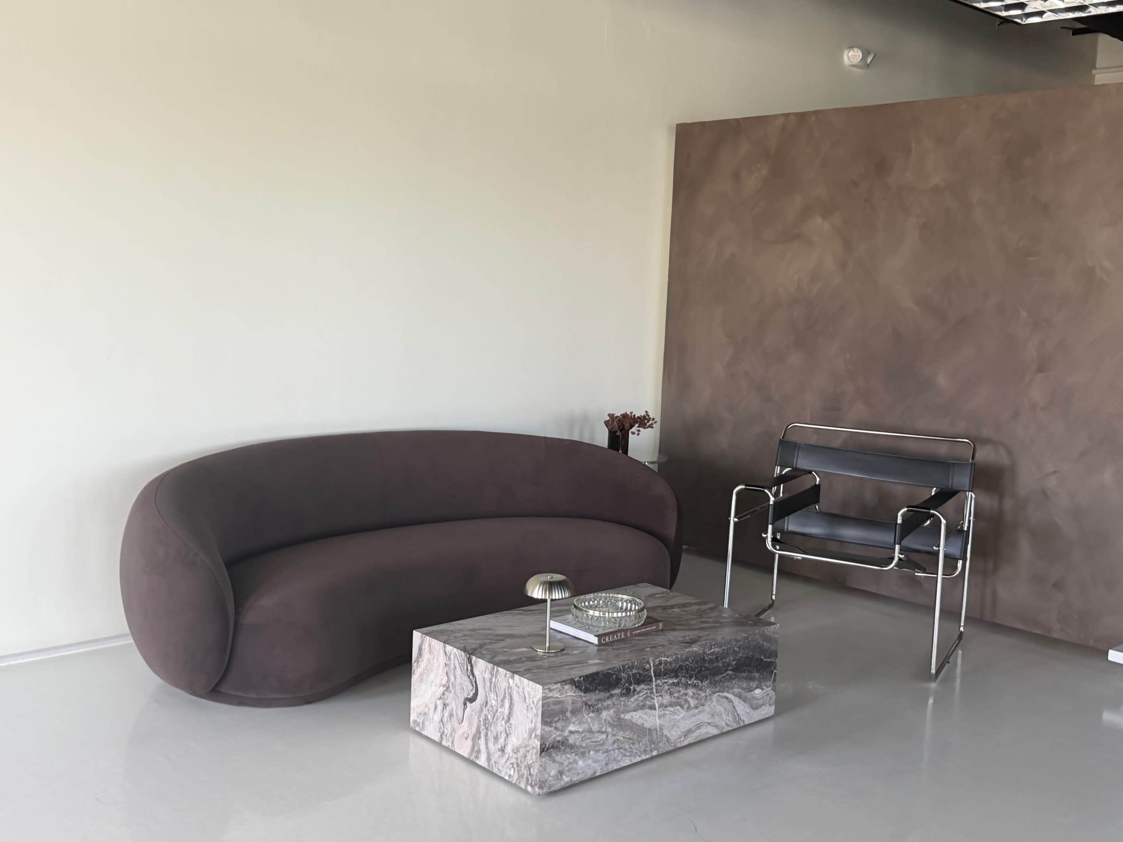 The image features a modern interior with a curved brown sofa, a marble coffee table, and a sleek metal chair against a light-colored wall.