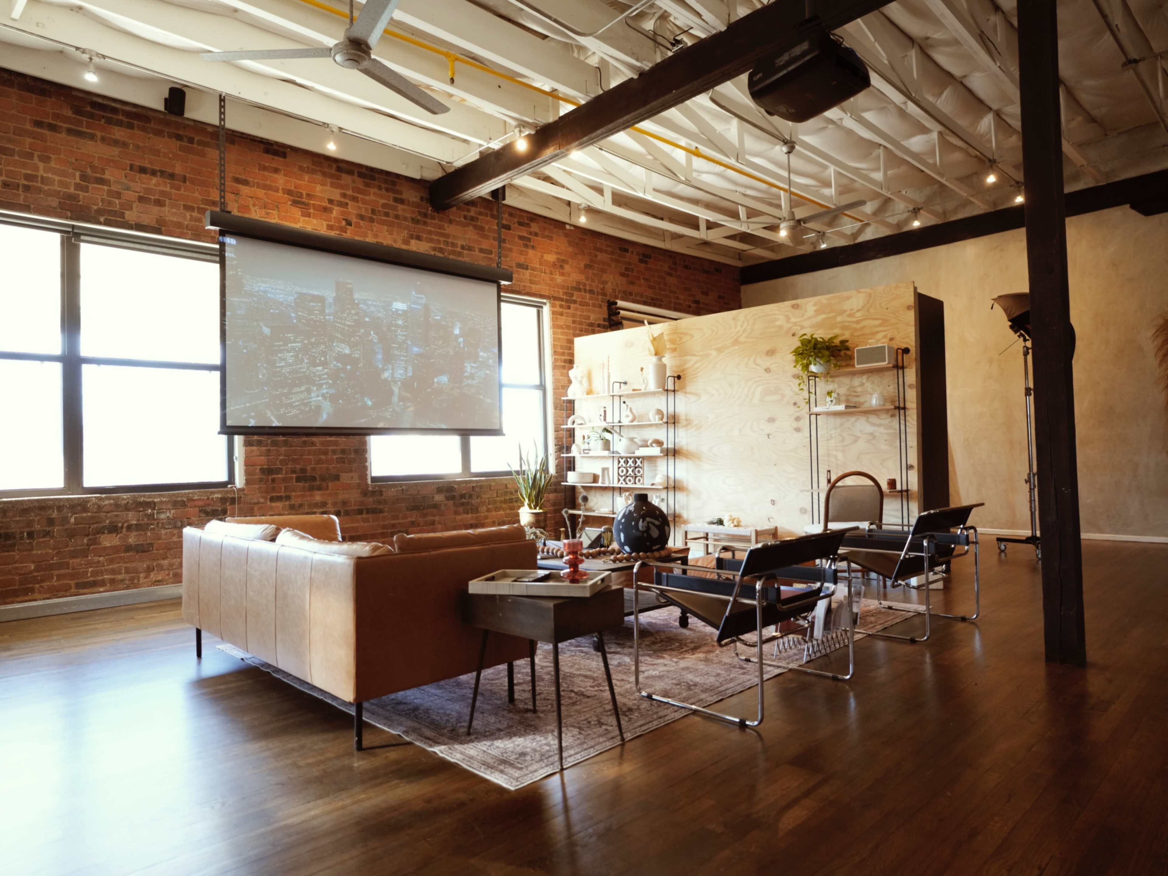 The image shows a modern living space with a large projection screen, a brown leather sofa, and various seating arrangements around a coffee table, set against a backdrop of exposed brick walls.