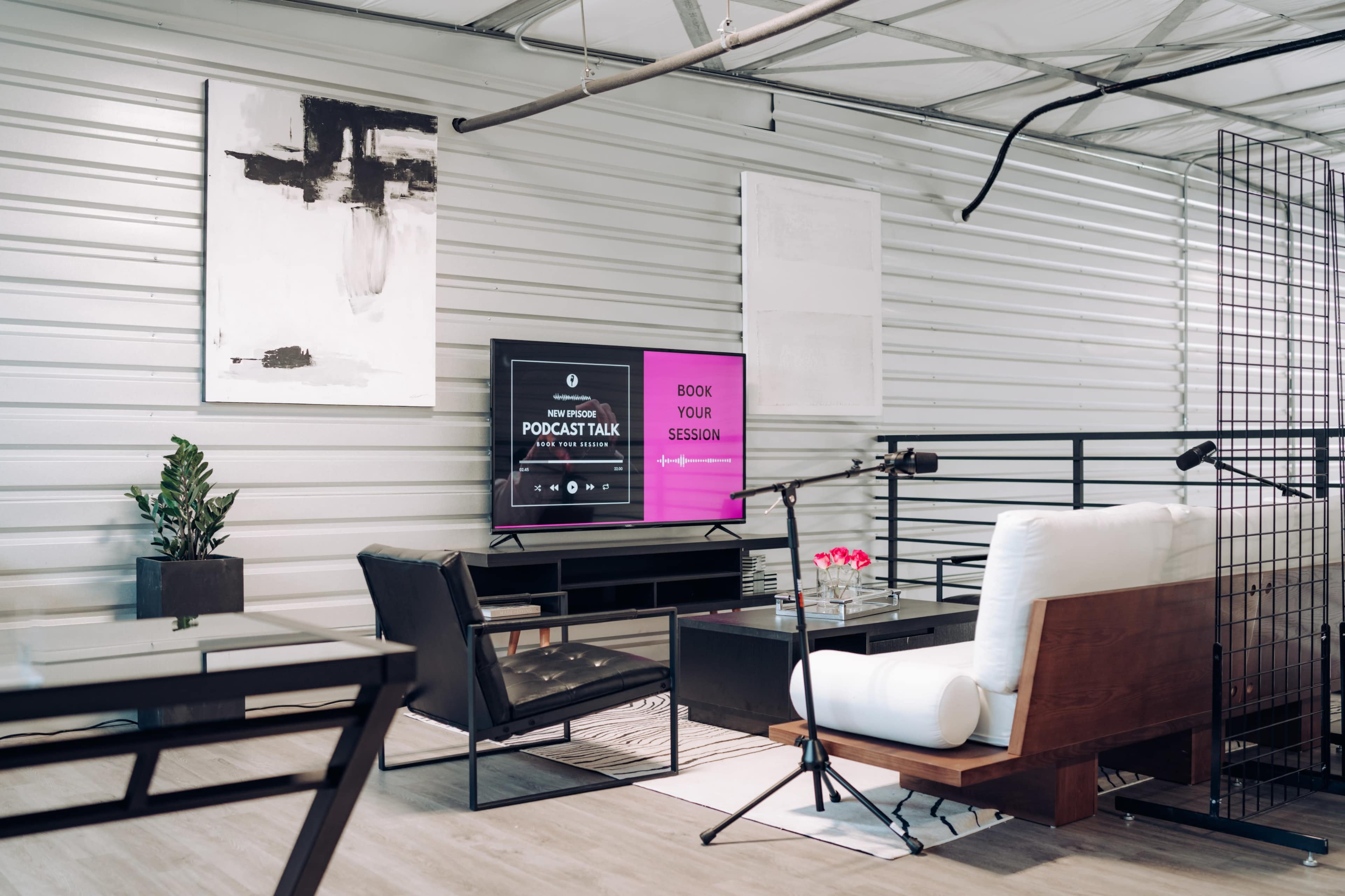 A modern podcasting space features a television displaying a booking interface, a comfortable seating area, and microphones set up for recording.