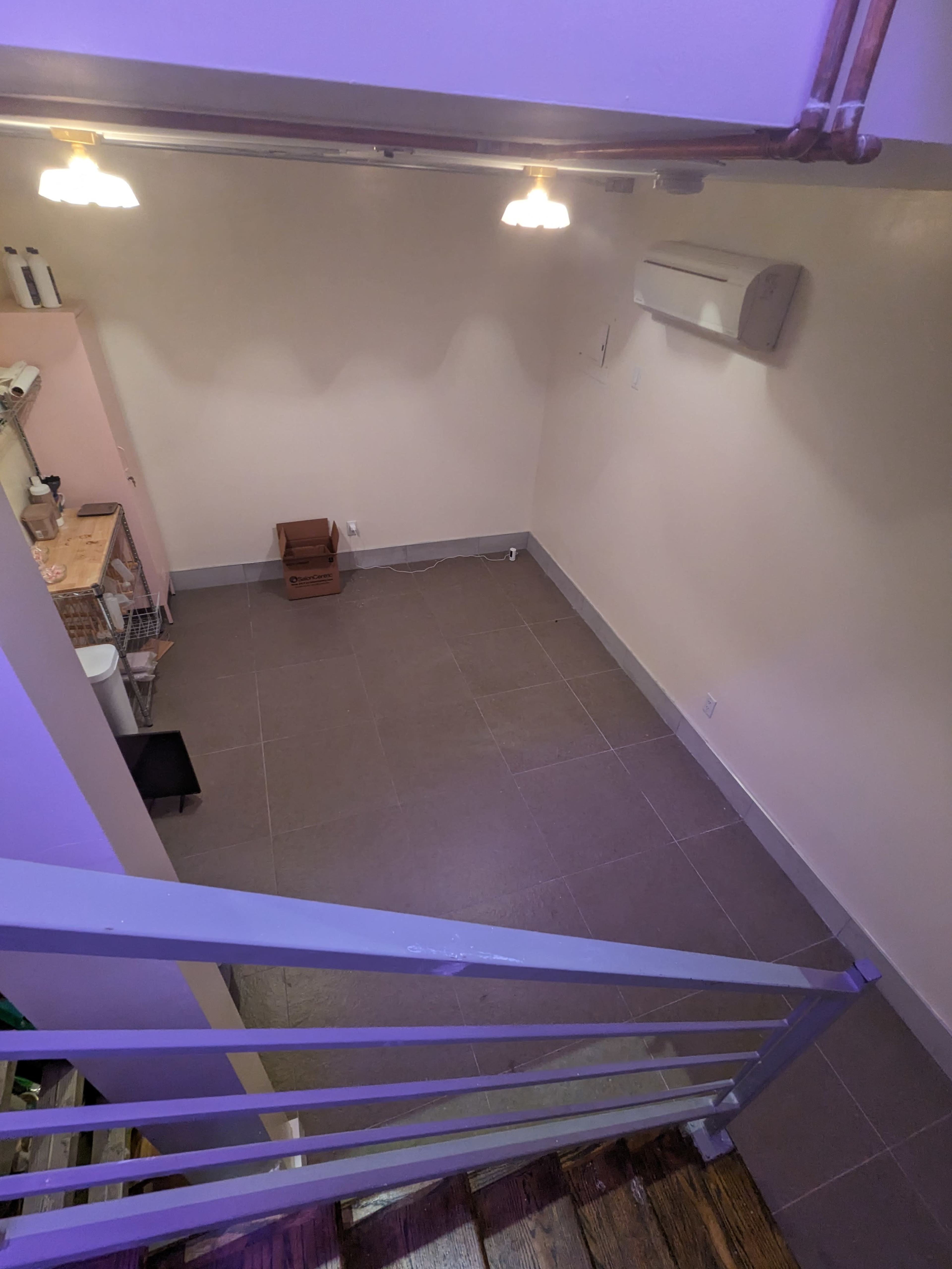 A view from above shows a small, empty room with tiled flooring, pink walls, and a kitchenette along one side.