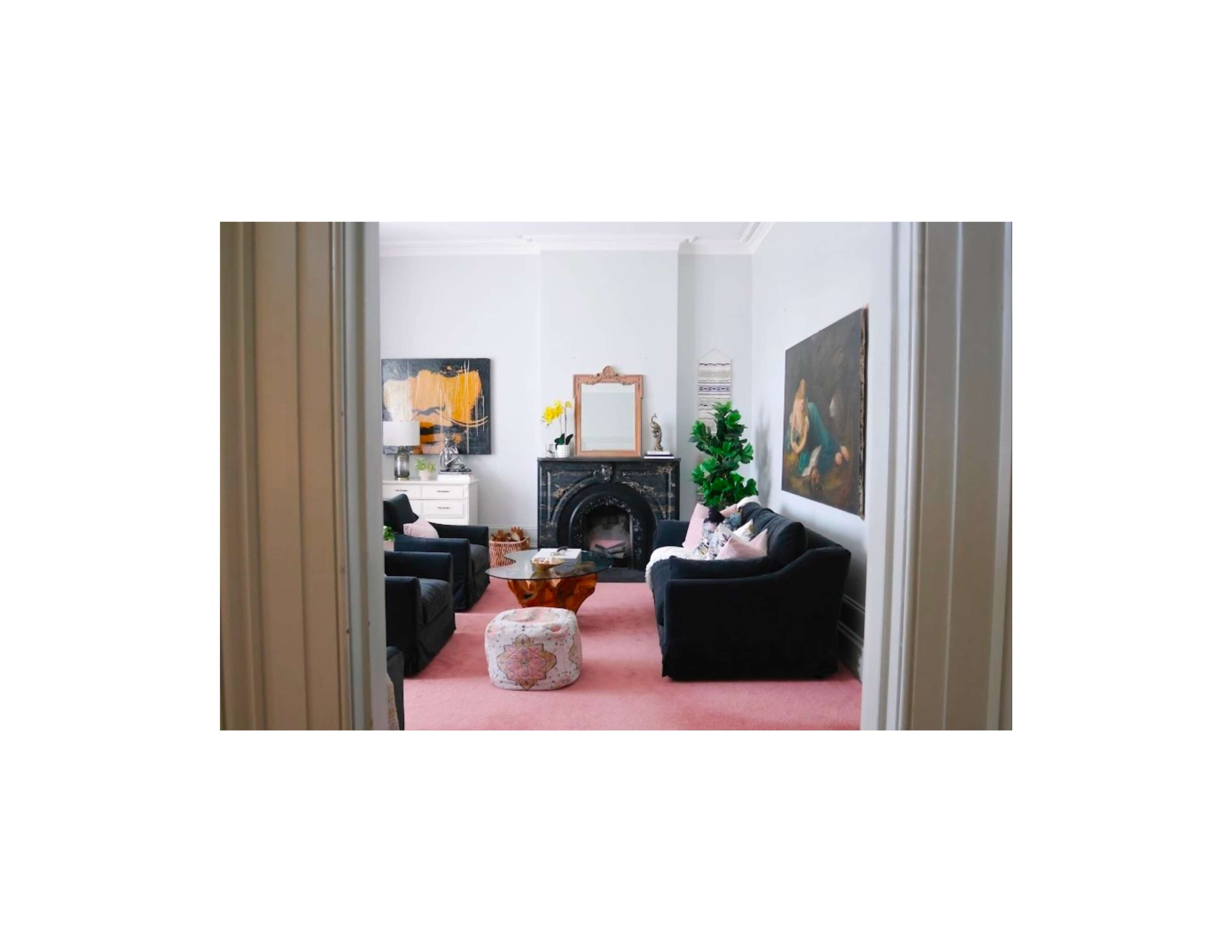 A living room features a fireplace, two dark sofas, a pink rug, and several pieces of wall art.