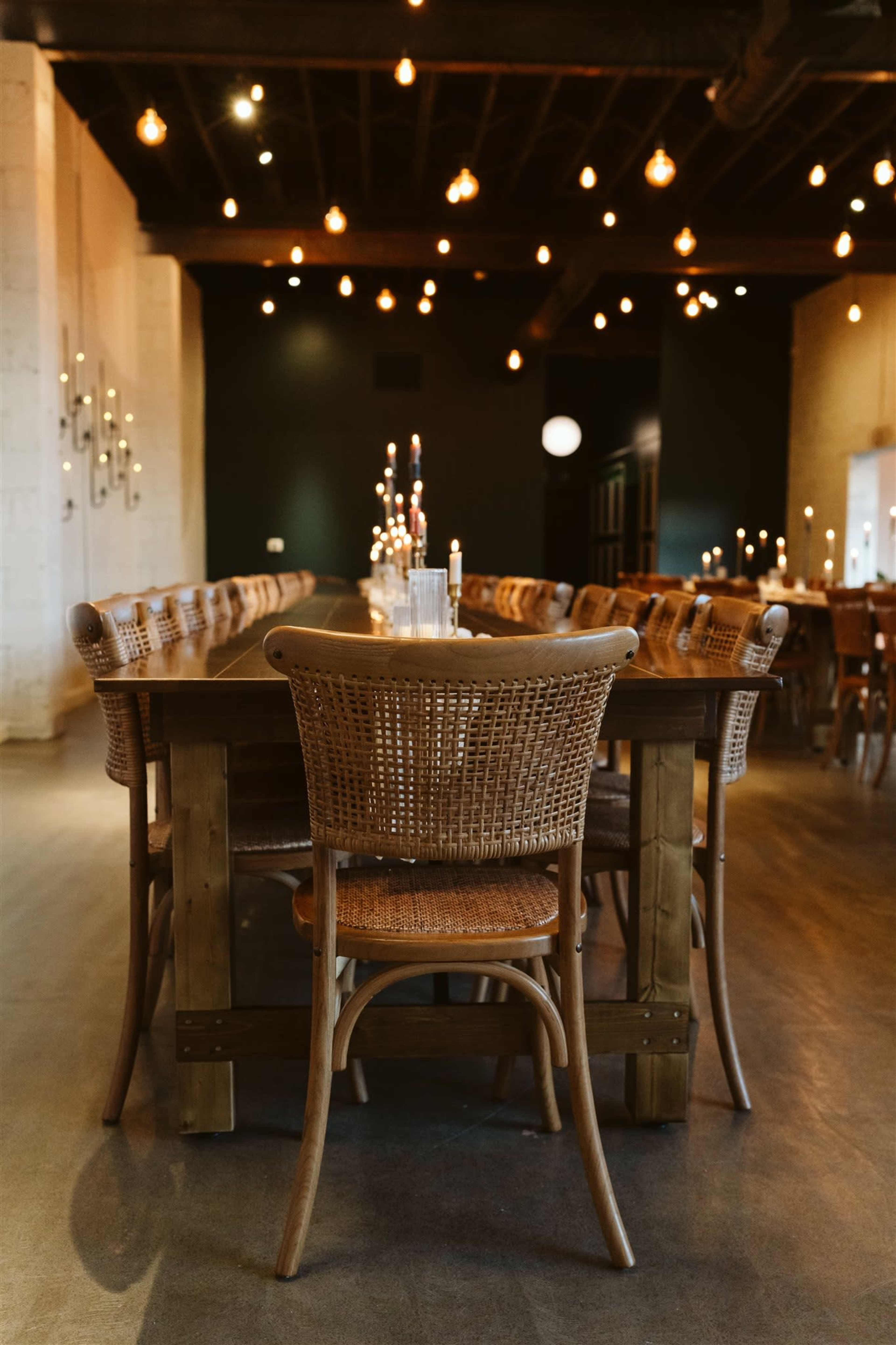 A long wooden table surrounded by woven chairs is set in a dimly lit restaurant with hanging light bulbs above.