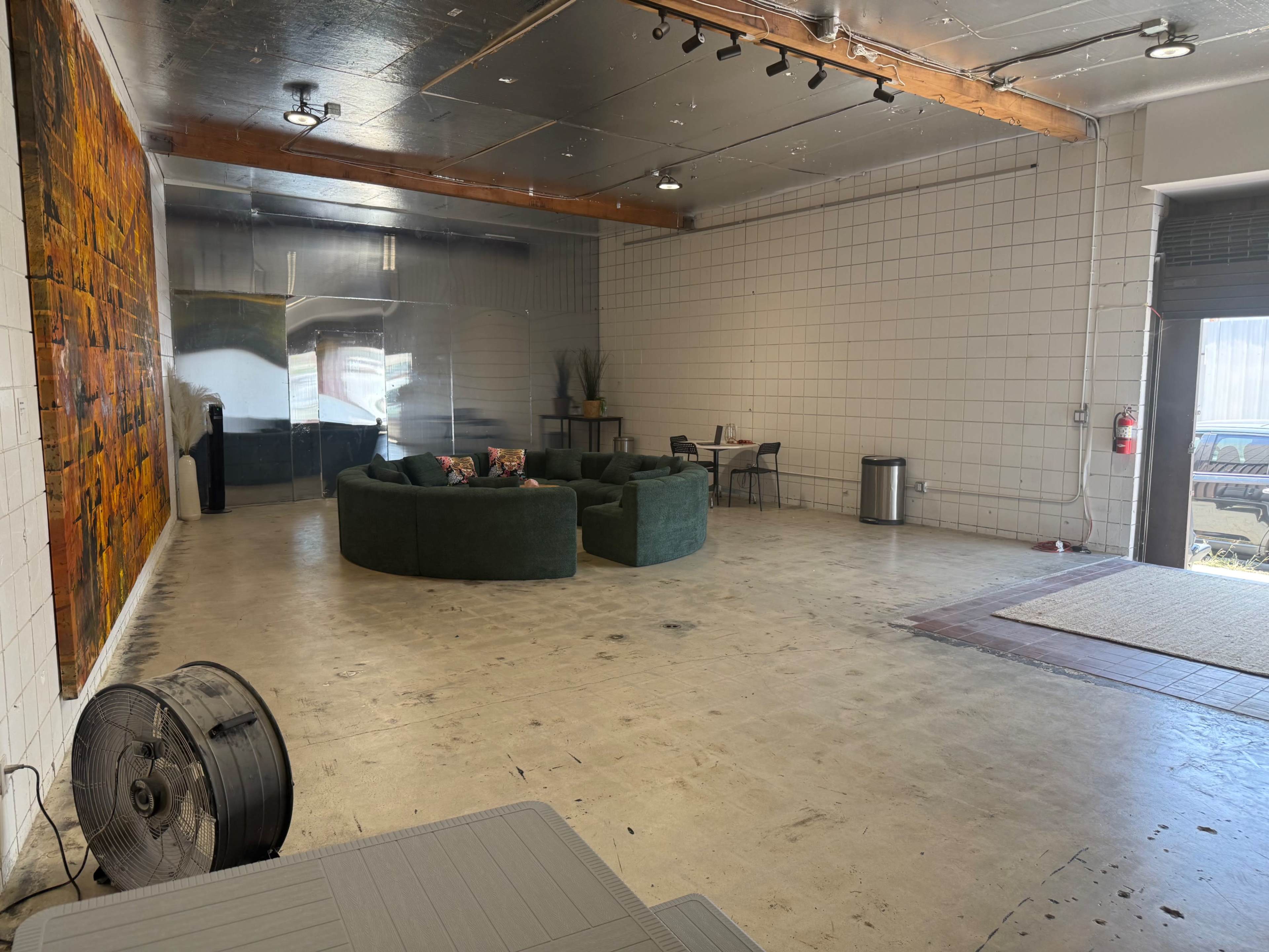 The image shows a spacious, empty room featuring a large green sectional sofa, a small dining table, and a fan on the floor, with a textured wall art piece visible in the background.