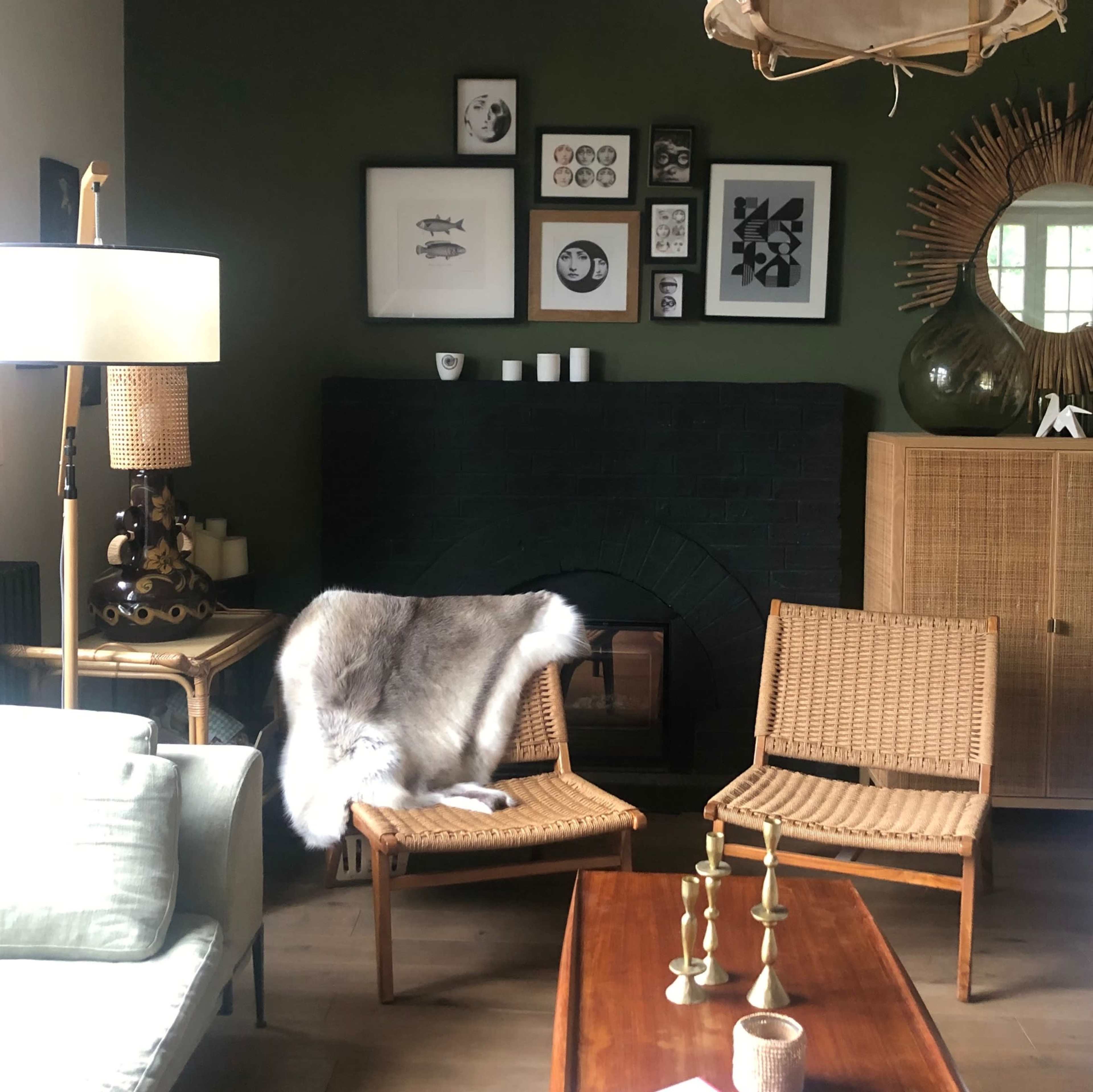 The image shows a cozy living room with a dark green wall, a black fireplace, wicker chairs, a wooden coffee table, and various framed artworks on display.
