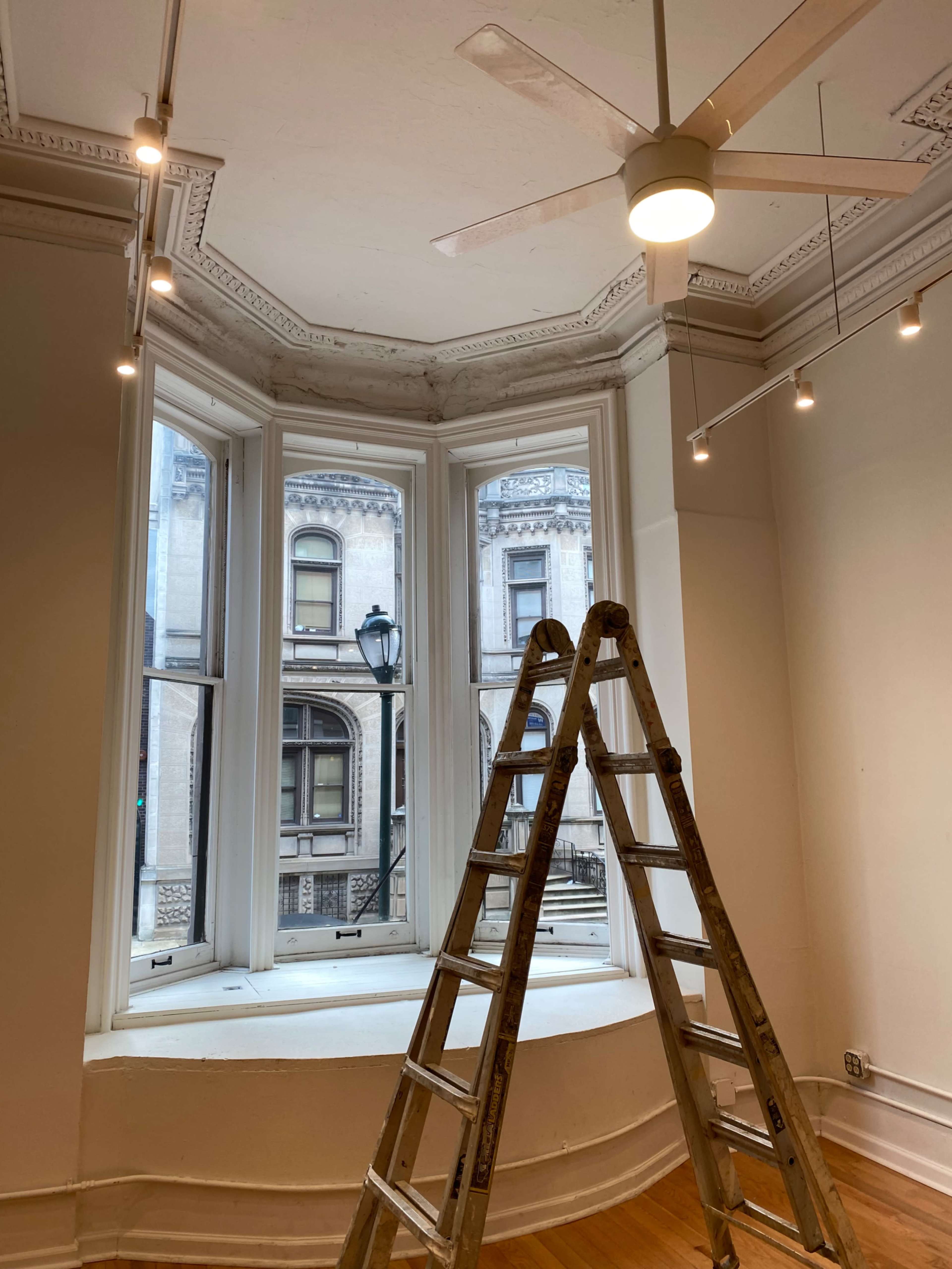 A wooden ladder stands in front of a large bay window with overhead lights and a view of an adjacent building outside.