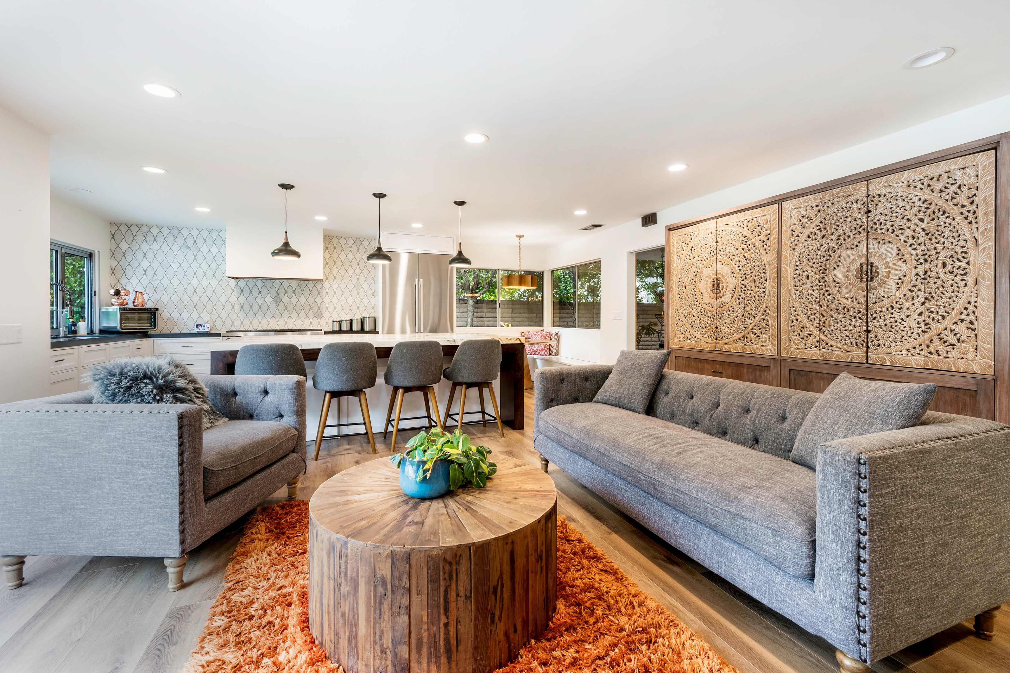 A modern living space features a gray sofa, a round wooden coffee table, and a dining area with bar stools near a kitchen with patterned backsplash.