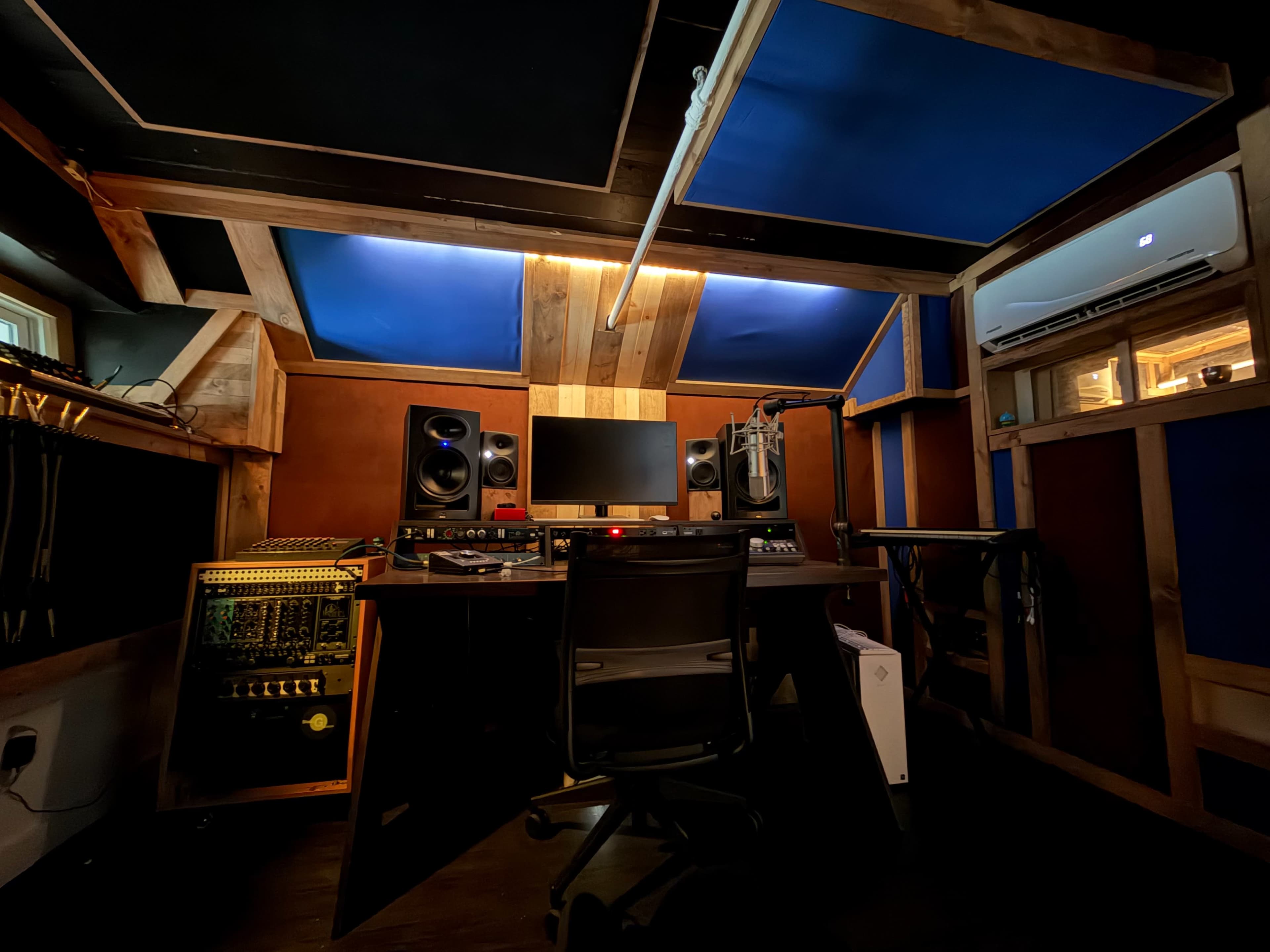 The image shows a recording studio with a central workstation, equipped with speakers, a computer monitor, and various audio equipment, all surrounded by wooden walls and a blue ceiling.
