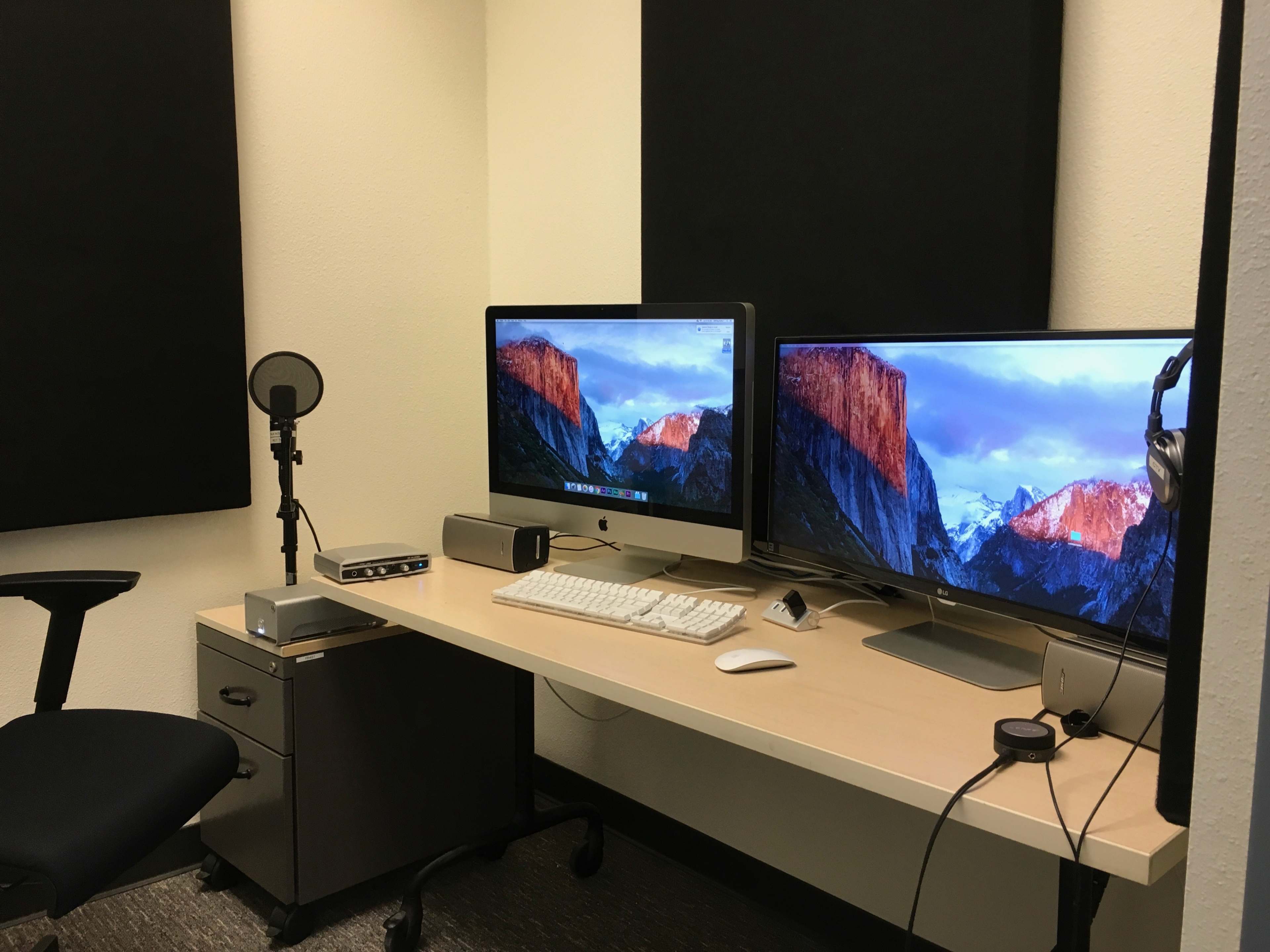 The image shows a compact office space with two monitors, a microphone, and a keyboard placed on a desk, alongside a small filing cabinet and headphones.