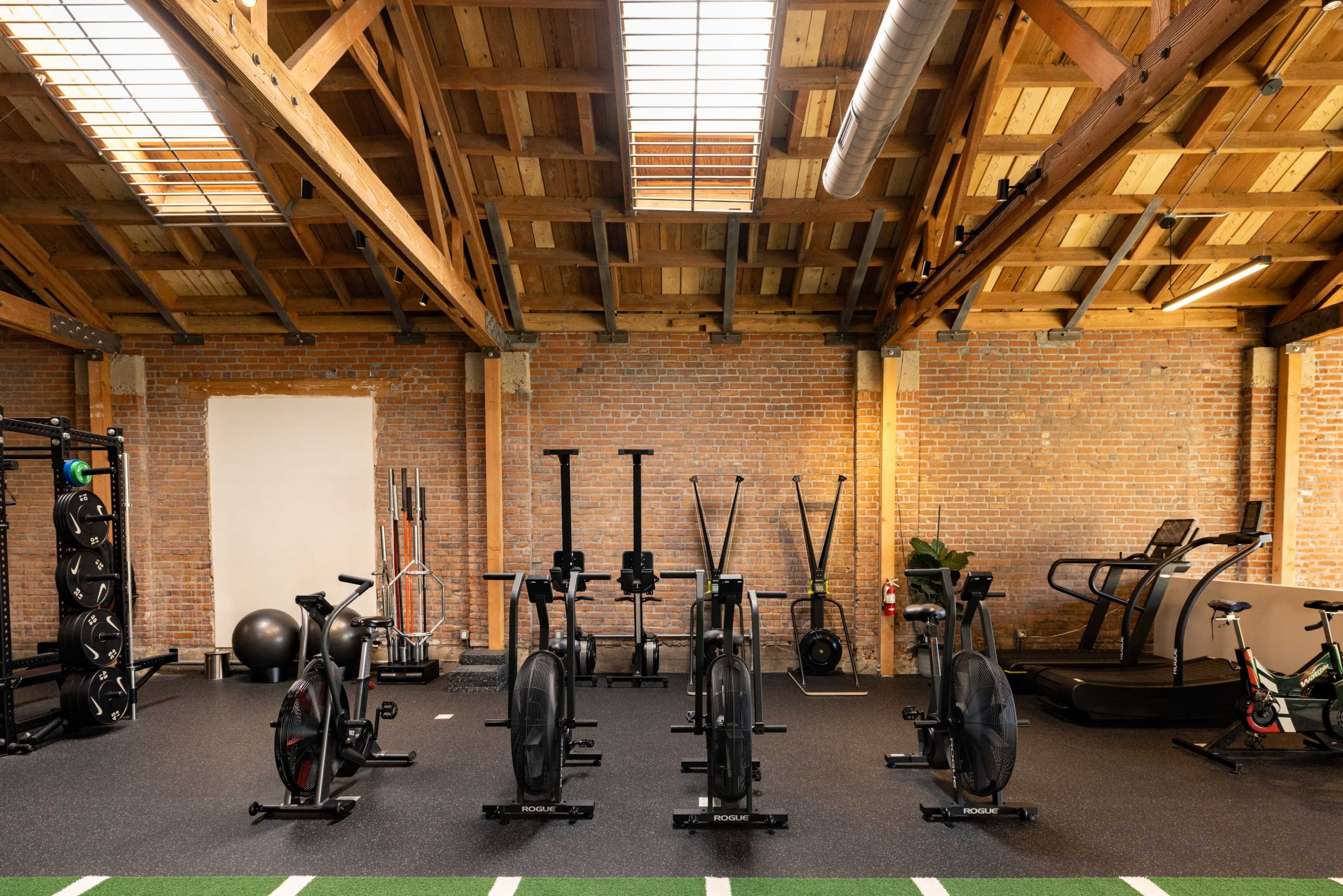 The image shows a spacious gym with a variety of exercise equipment lined up against a brick wall and under a wooden ceiling.