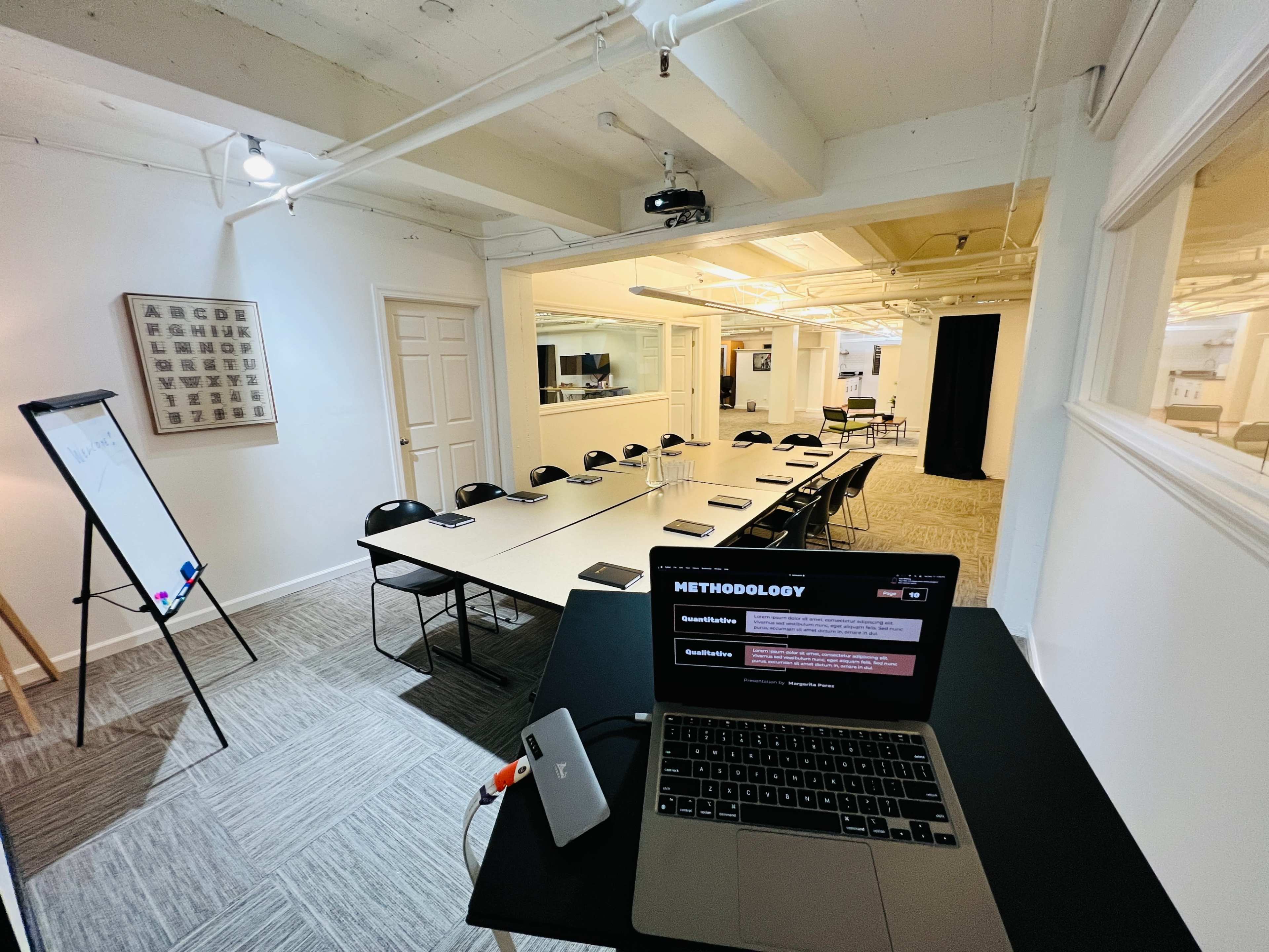 A conference room with a long table set for a meeting, featuring a laptop on a side table and a whiteboard in the foreground.