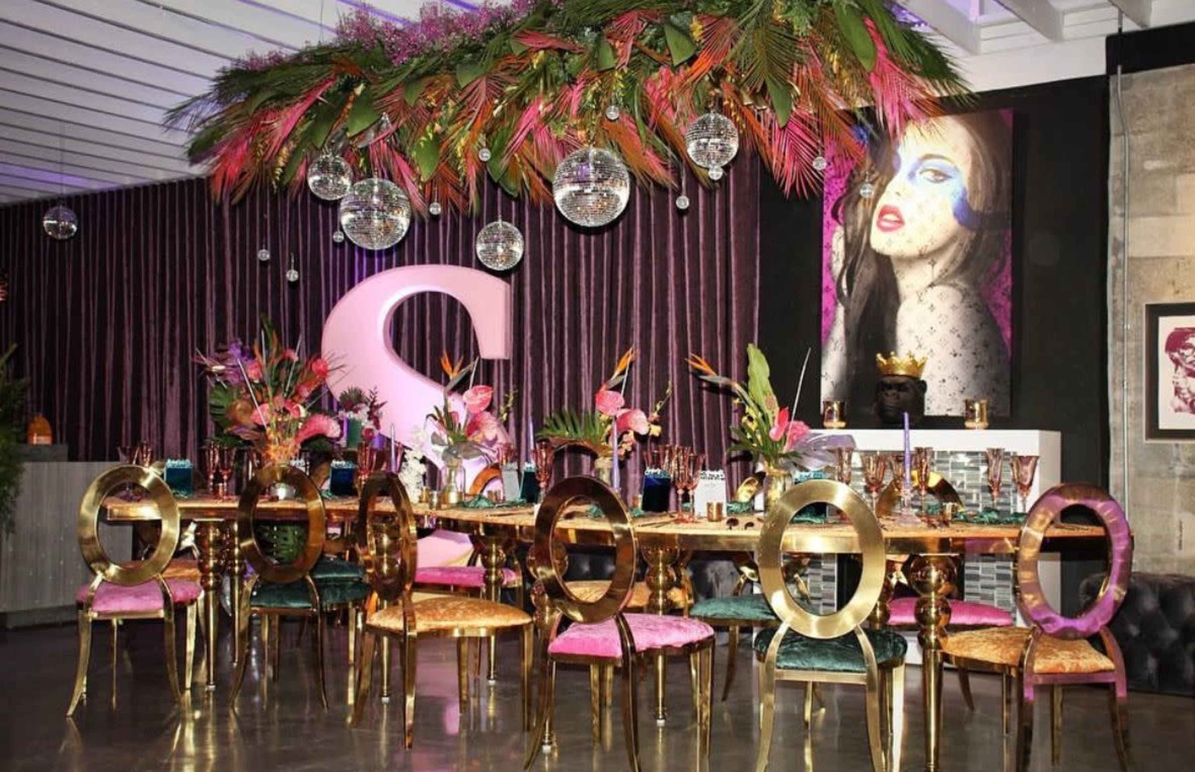 A lavish dining setup features a long table with ornate gold chairs, colorful floral arrangements, and disco balls hanging from a vibrant overhead display.