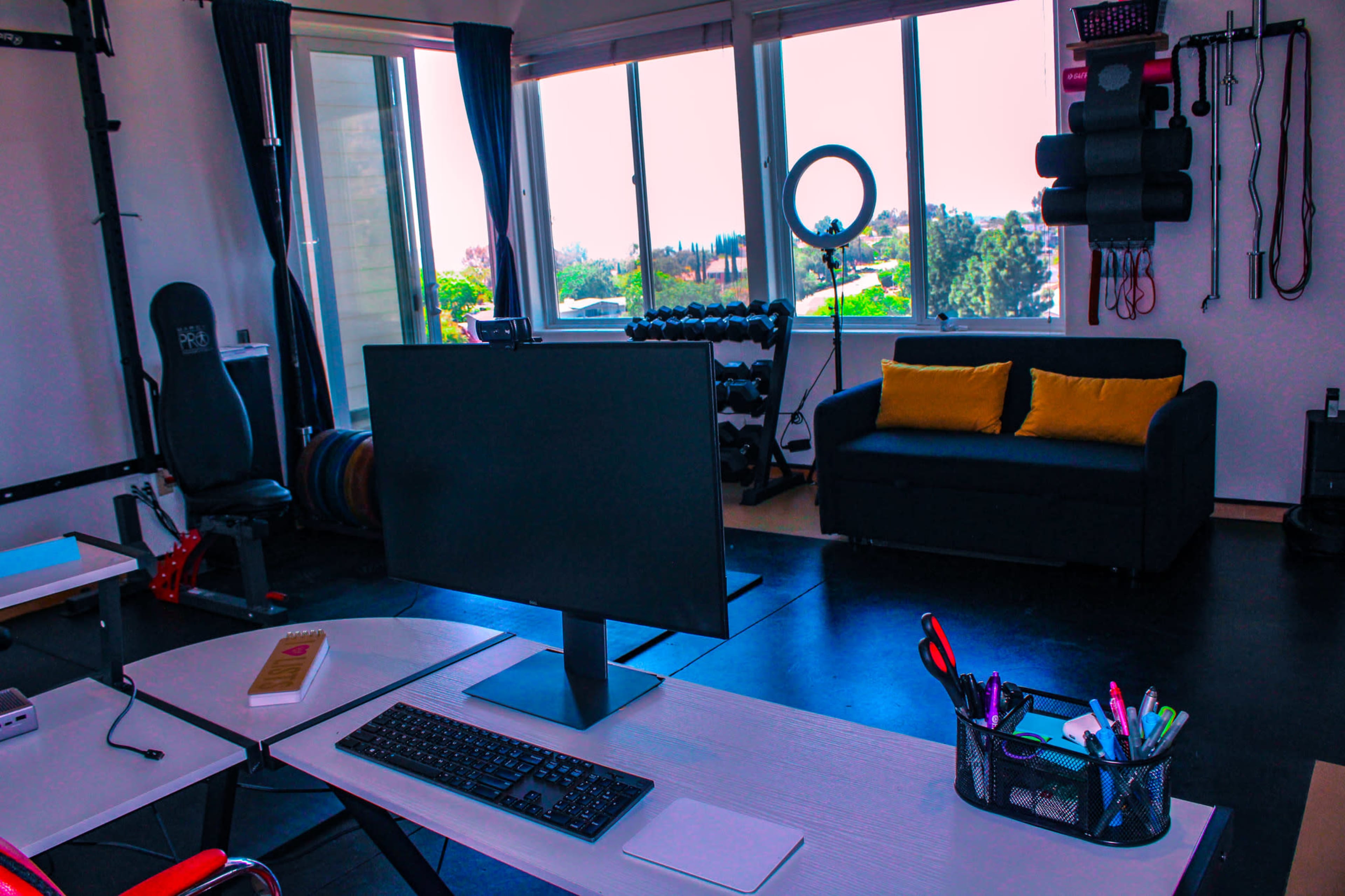 A home office space with a computer setup on a desk, a black couch with yellow pillows, and workout equipment visible through large windows.