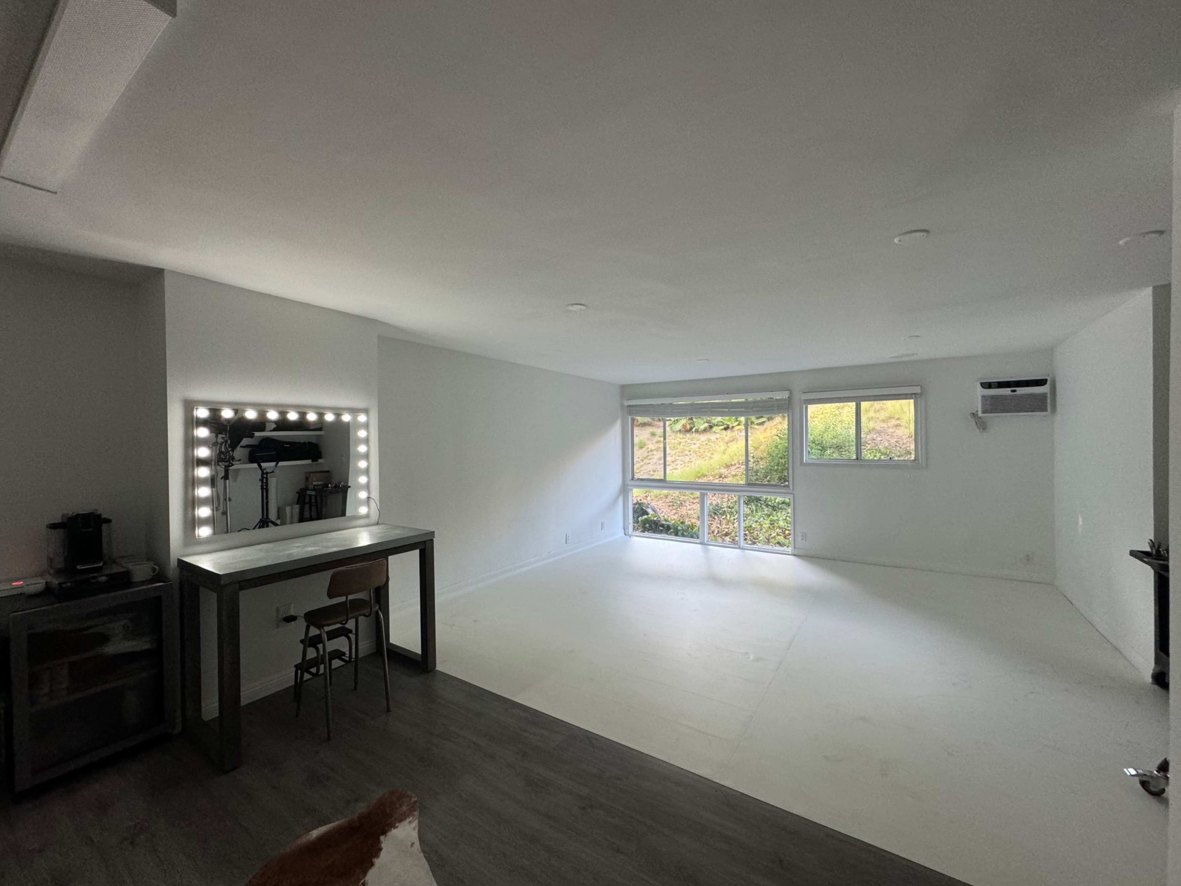 The image shows a spacious, empty room with large windows and a table with a mirror lined with lights.