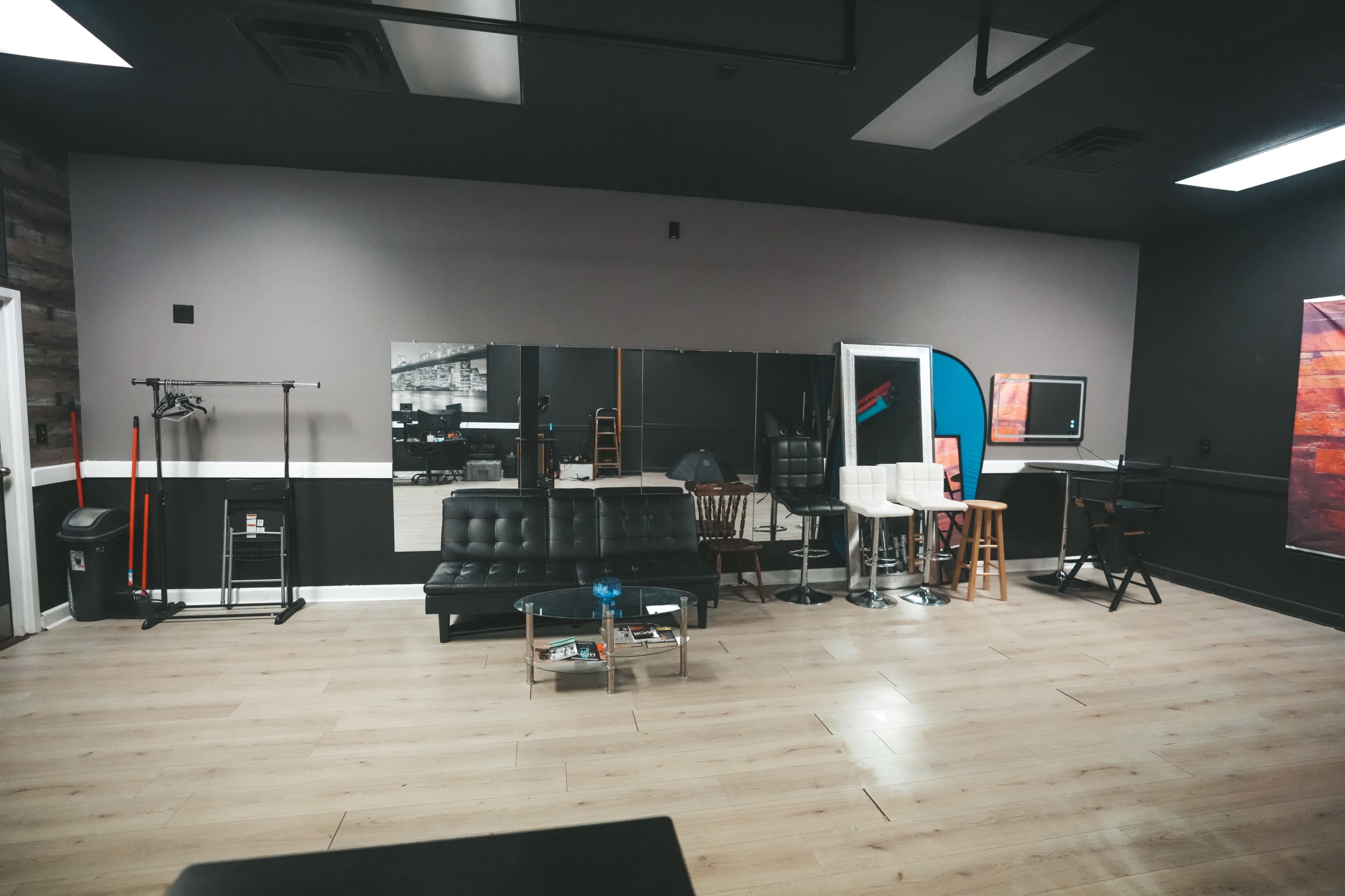 The image shows a spacious studio interior featuring a black leather sofa, a glass coffee table, various seating arrangements, and a large mirror on the wall.