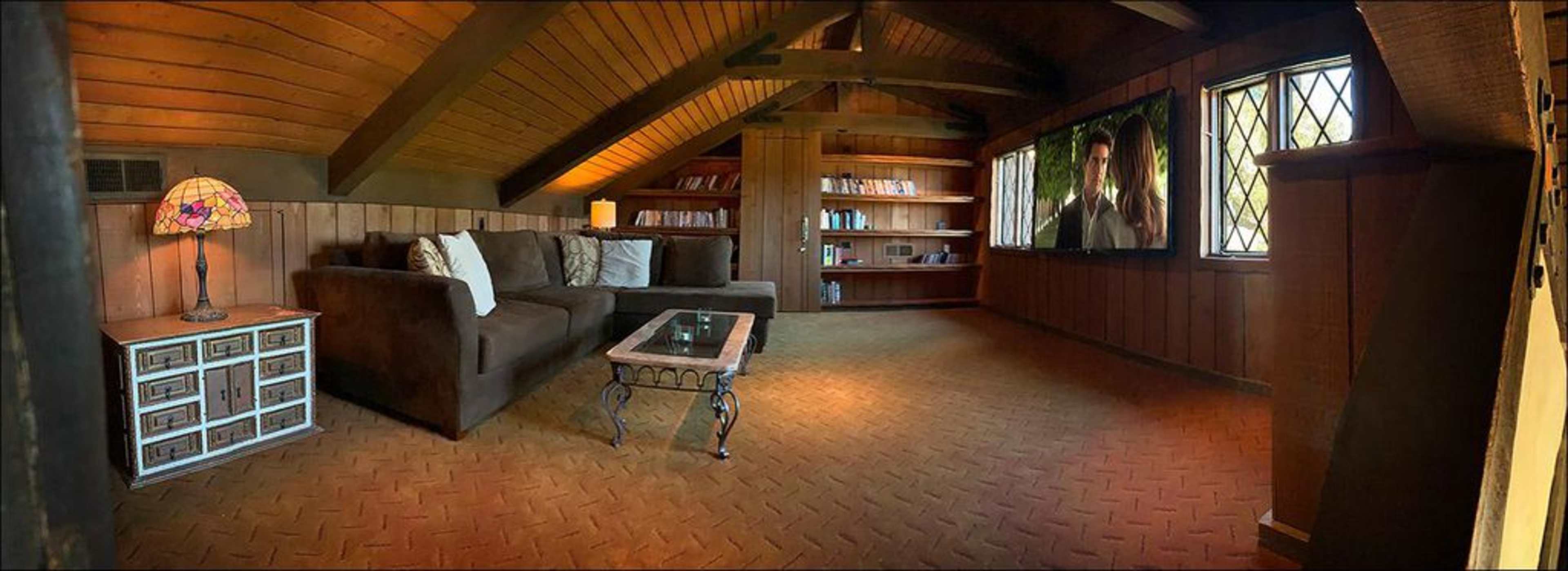 The interior of a cozy attic living space features a sectional sofa, a glass coffee table, a large television on the wall, and bookshelves lined with books.