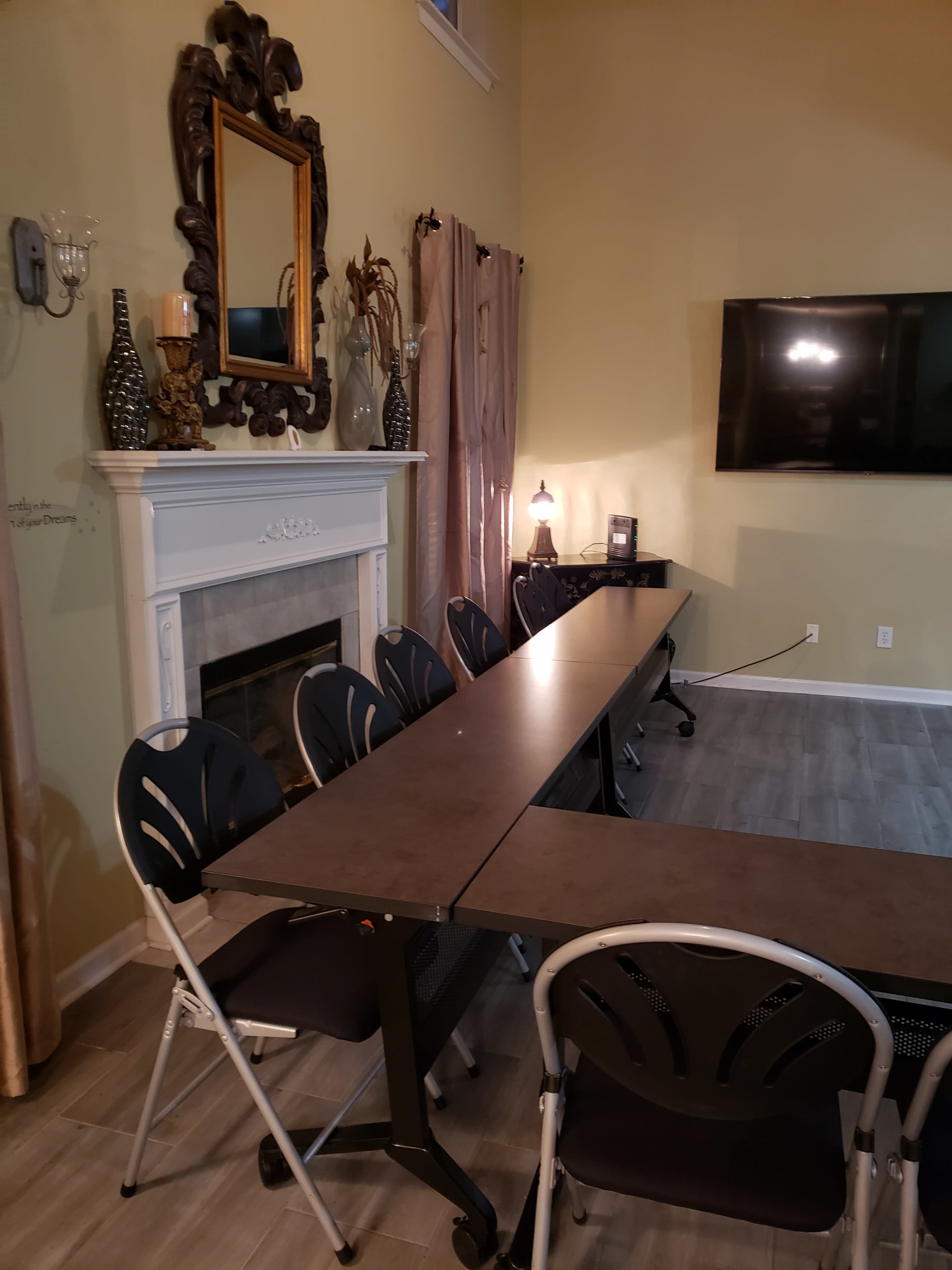 A rectangular table with folding chairs is set up in a room featuring a fireplace, a mirror, and a television mounted on the wall.