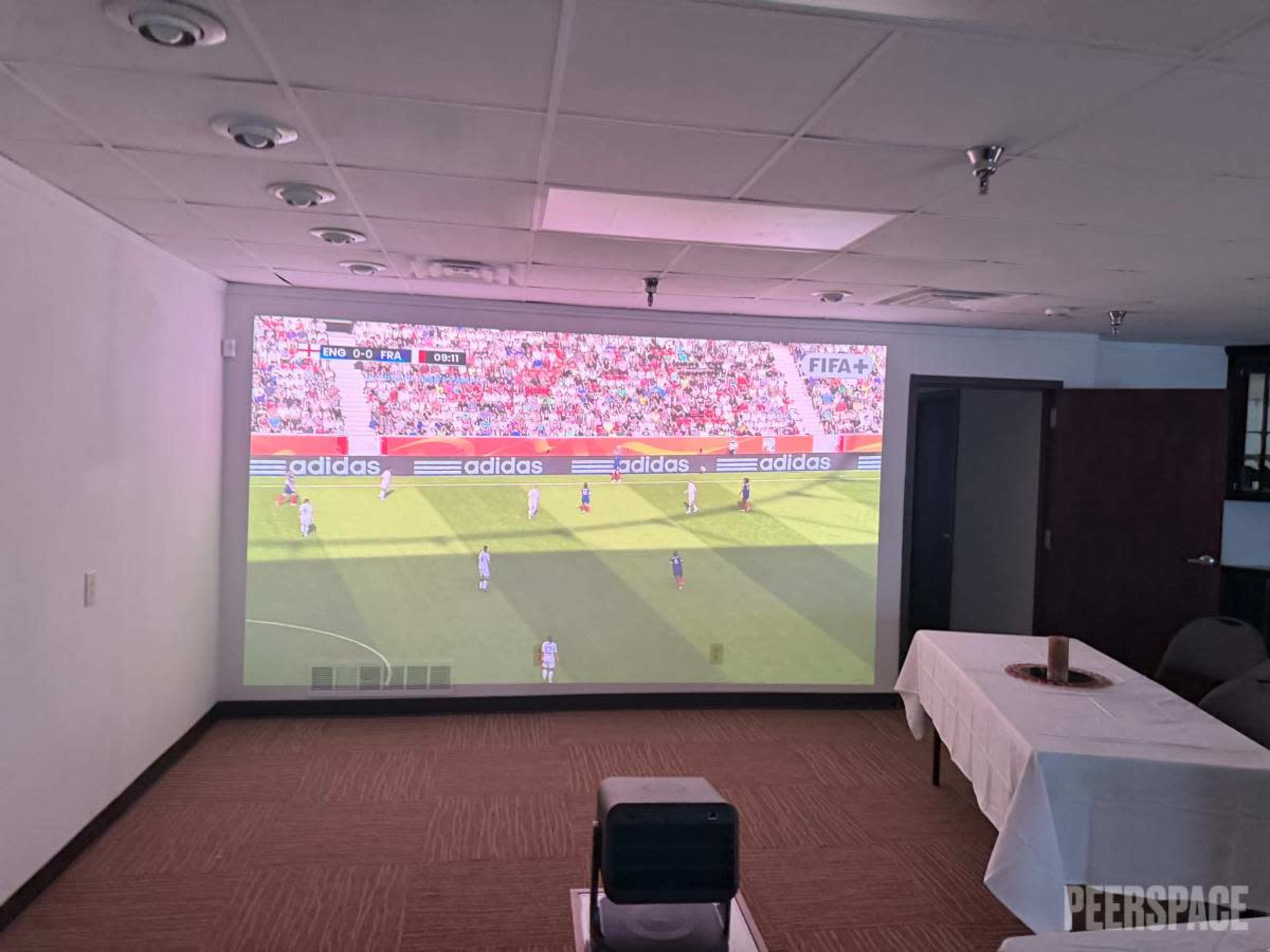 A large projector screen displays a soccer match in a conference room with a table and chairs in the foreground.