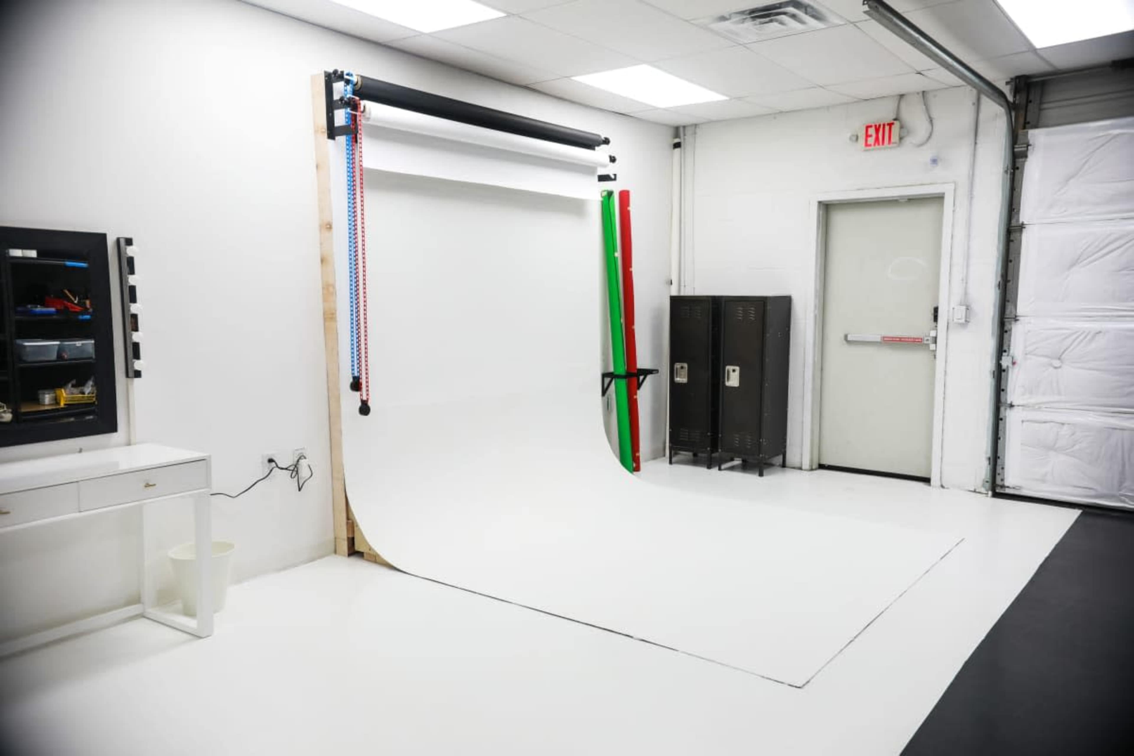 The image shows a photography studio with a white backdrop, lighting equipment, and storage lockers against the wall.