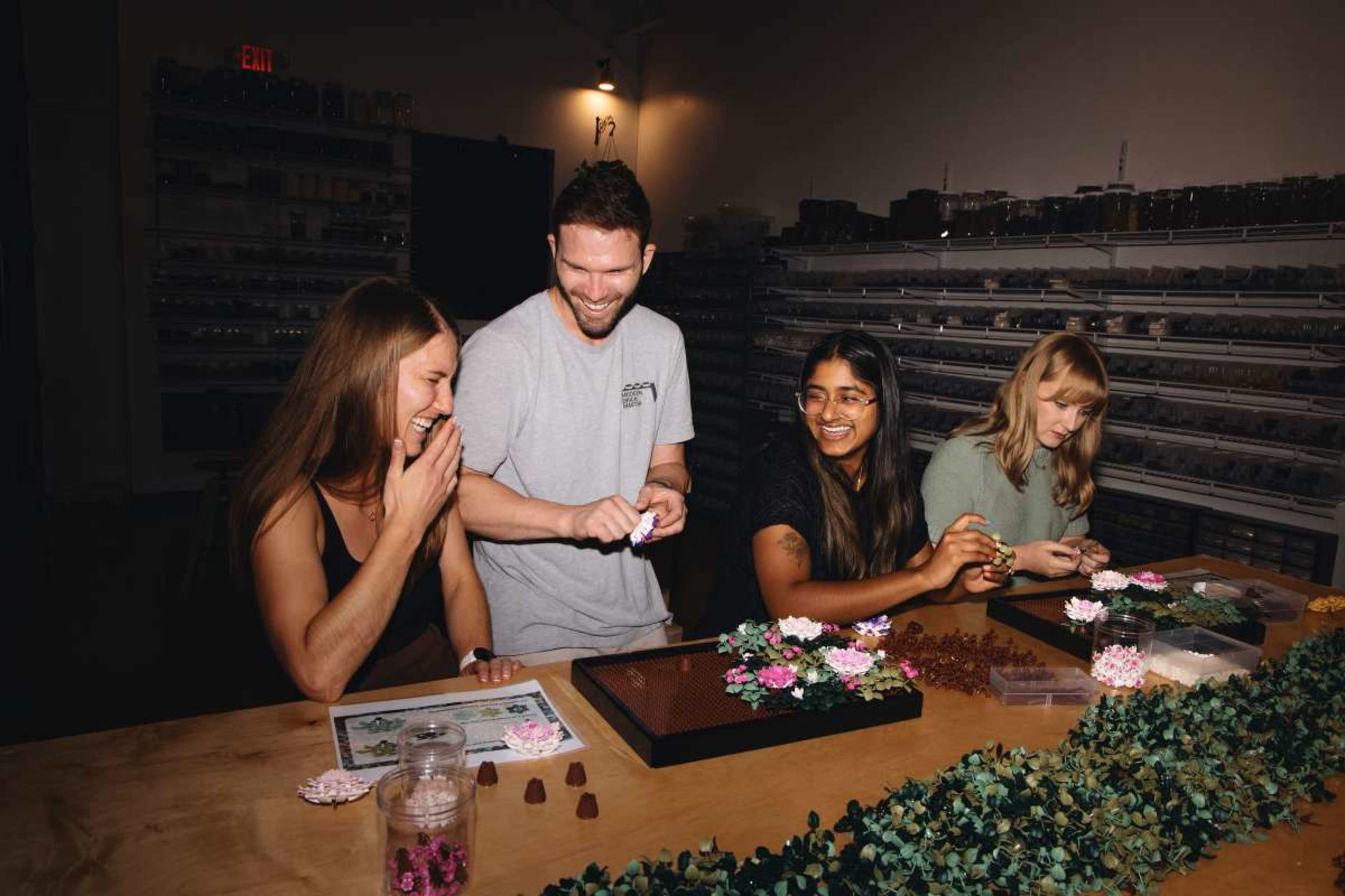 A group of four people is seated around a table, engaged in crafting with flowers and various materials.