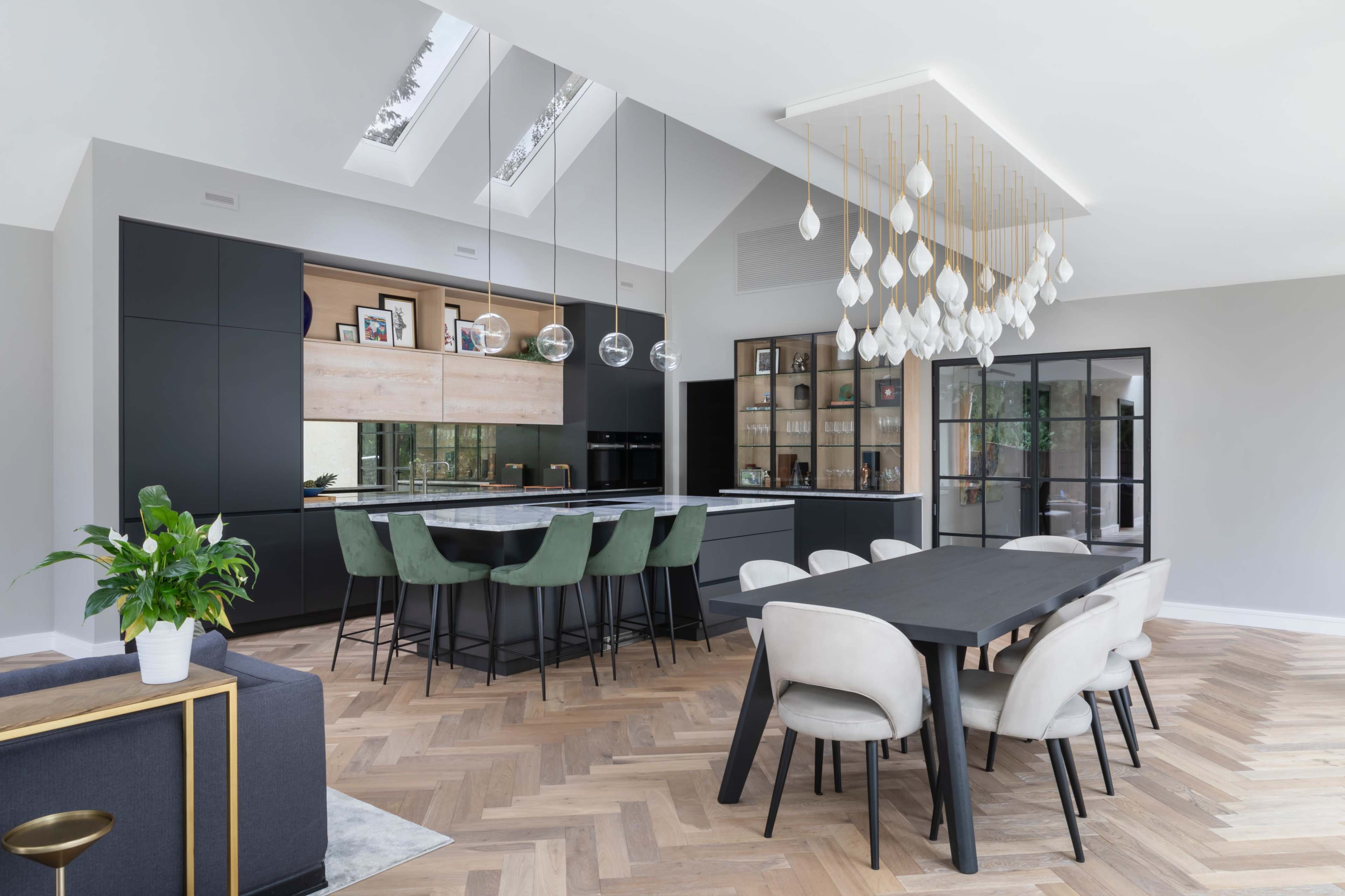 A modern kitchen and dining area with a large, light wooden table, black cabinetry, and a distinctive pendant light fixture above the island.