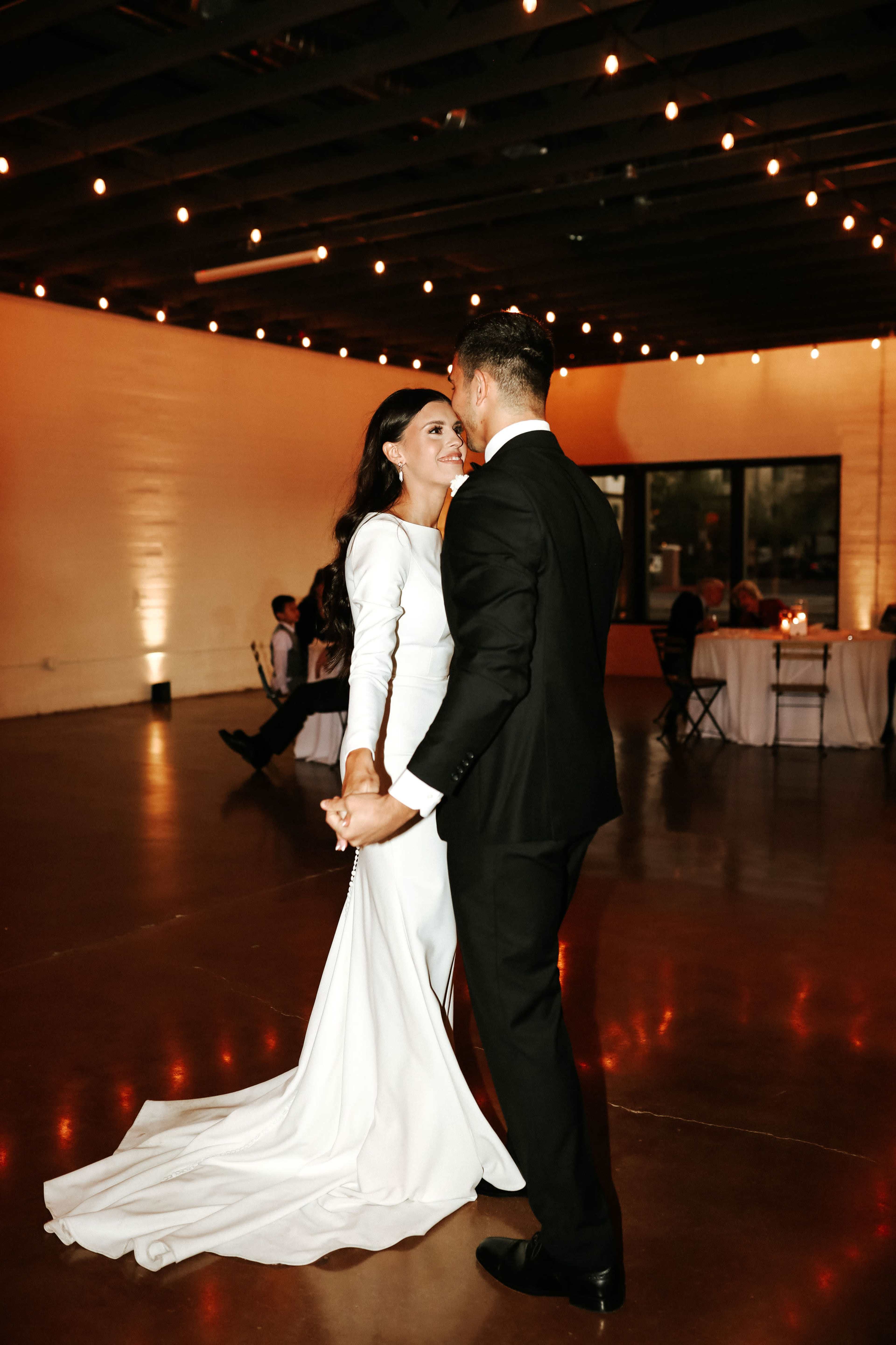 A couple dances closely in formal attire in a dimly lit event space adorned with string lights.